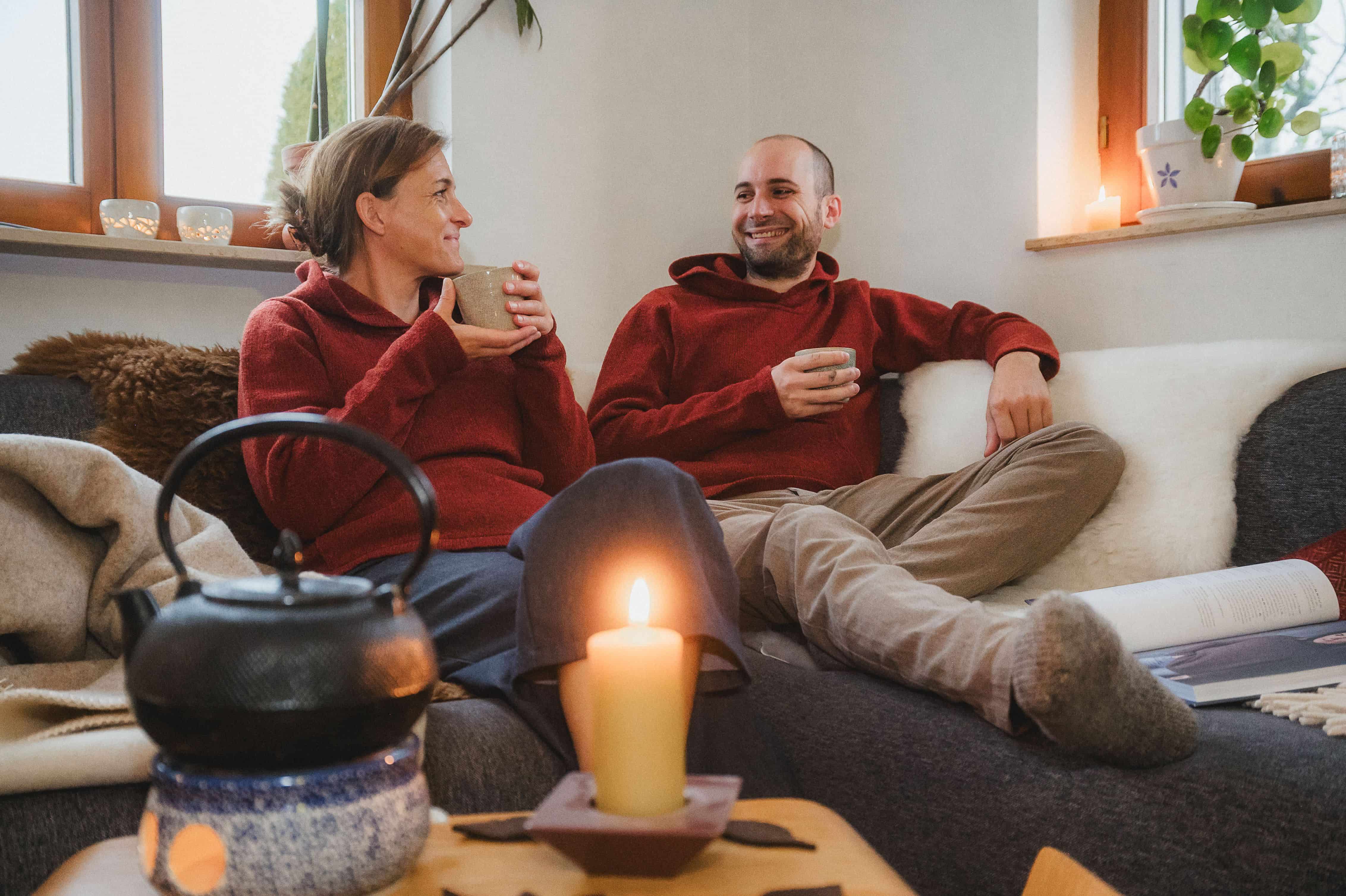 Mann und Frau mit roten Finkhof Kapuzenpullis aus bio Merinowolle (kbT) sitzen mit Tee auf einem Sofa