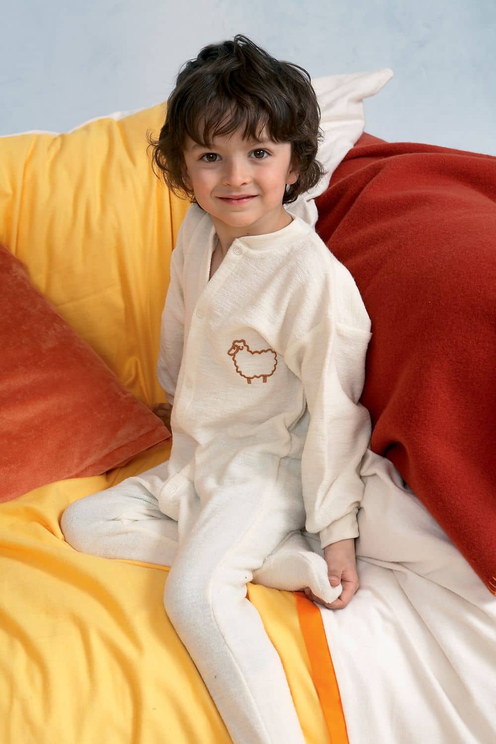 Einteiliger Schlafanzug naturweiß Kind sitzt mit hellem einteiligen Schlafanzug aus bio Wolle (kbT) mit Schaf auf der Brust auf Decken und Kissen