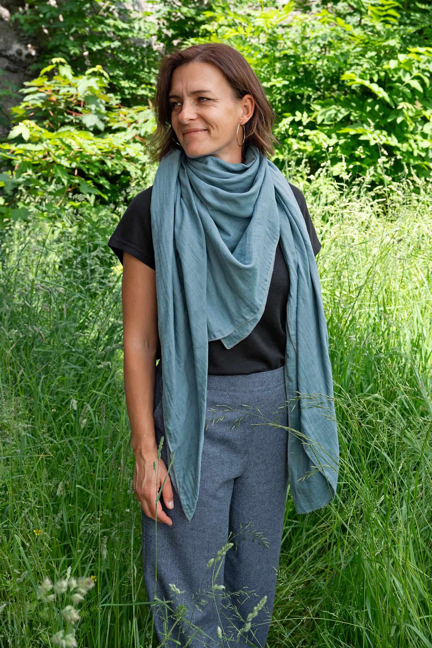 Frau steht auf Wiese und trägt eine blaue Hose, ein schwarzes Shirt und einen Schal aus blauem Baumwollmusselinstoff