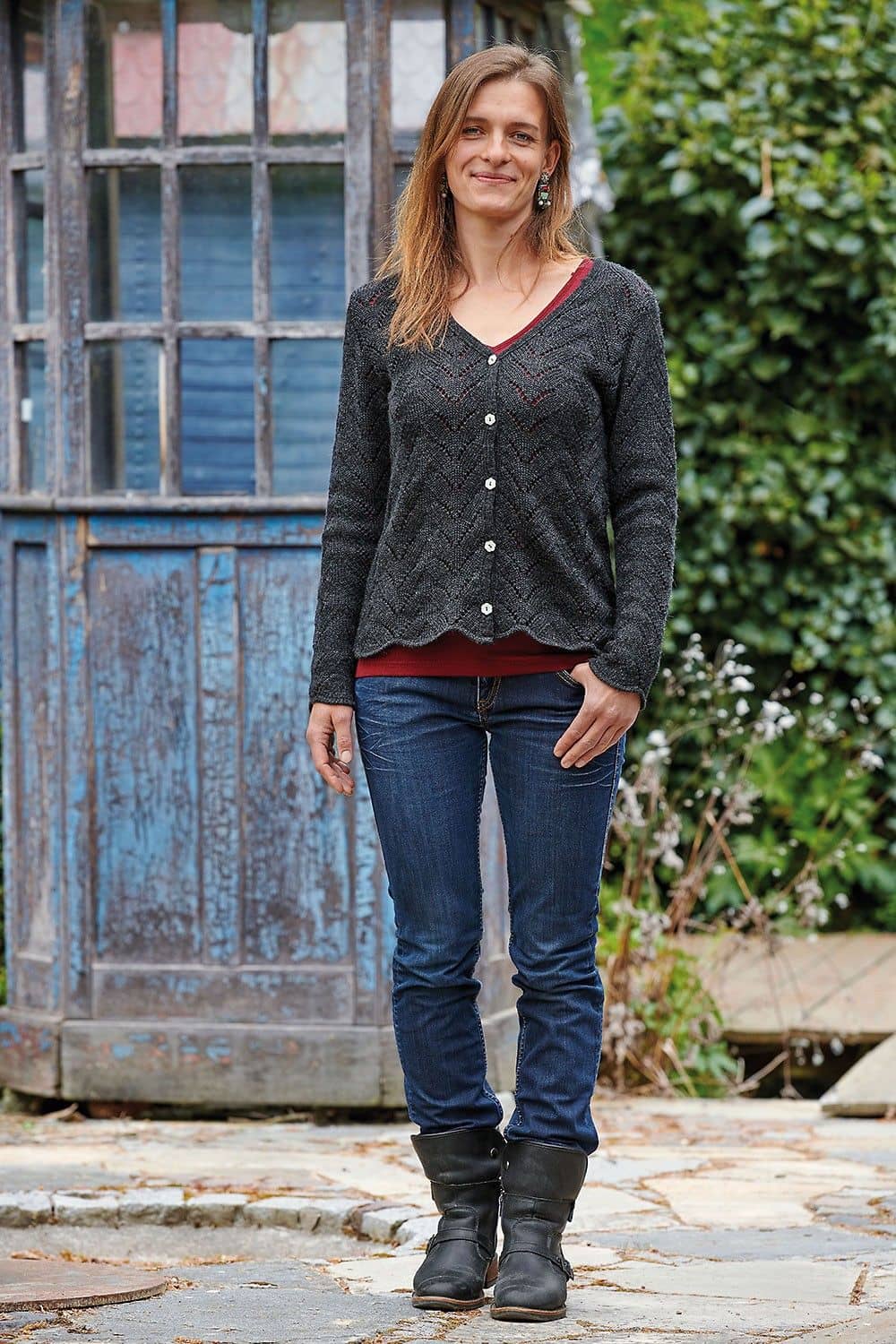 Frau trägt graue Ajourjacke aus bio Wolle (kbT) mit rotem Shirt, blauer Jeans und Stiefeln im Garten