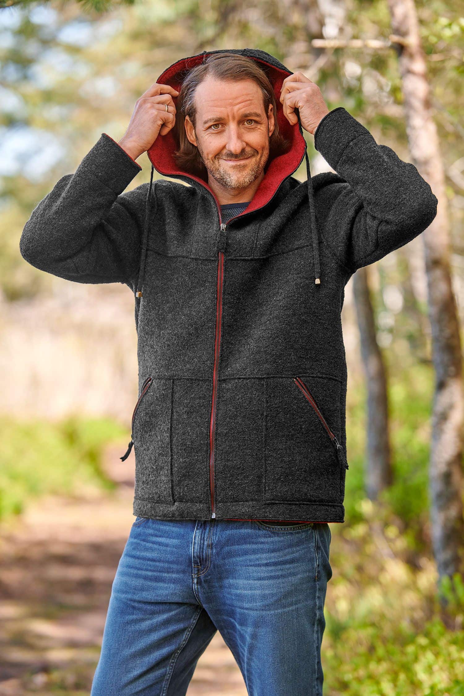 Mann mit Jeans setzt sich Kapuze von anthrazitgrauer Jacke mit roten Details aus bio Wolle (kbT) vom Finkhof auf