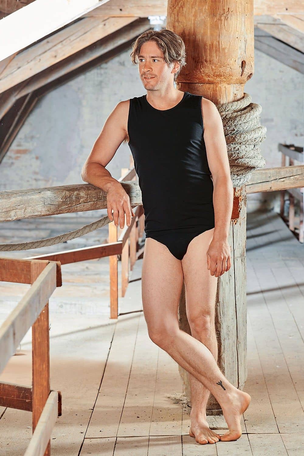 Mann mit schwarzem Slip aus bio Baumwolle (kbA) und Elasthan und schwarzem Top lehnt an Balken