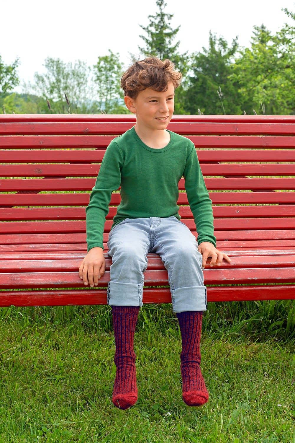 Junge sitzt auf roter Bank und trägt eine Jeans, ein grünes Shirt und rot/blaue Socken aus bio Wolle (kbT)