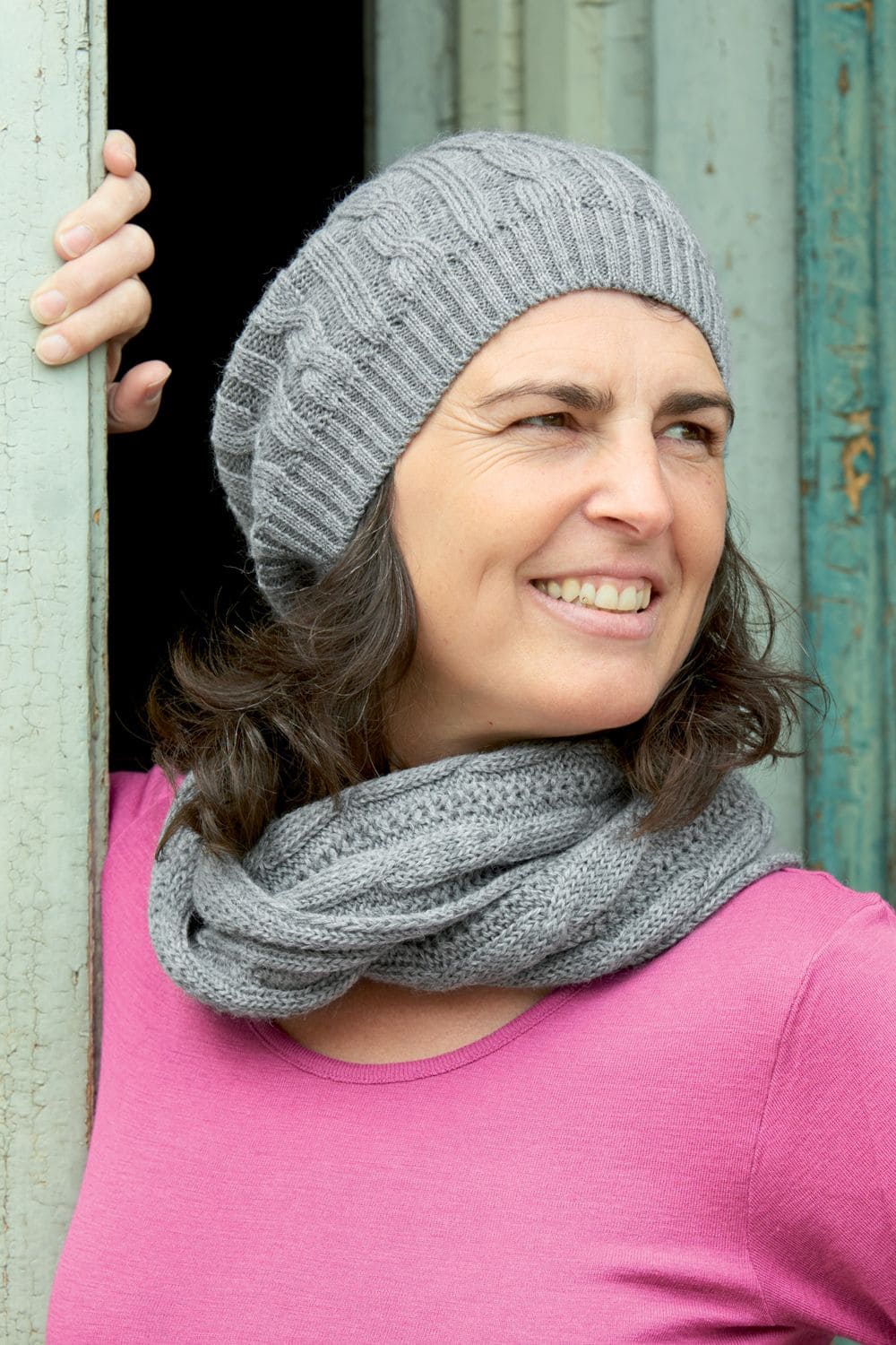 Frau trägt ein pinkes Shirt, graue Mütze und grauen Loopschal mit Zopfmuster aus bio Wolle (kbT) vom Finkhof