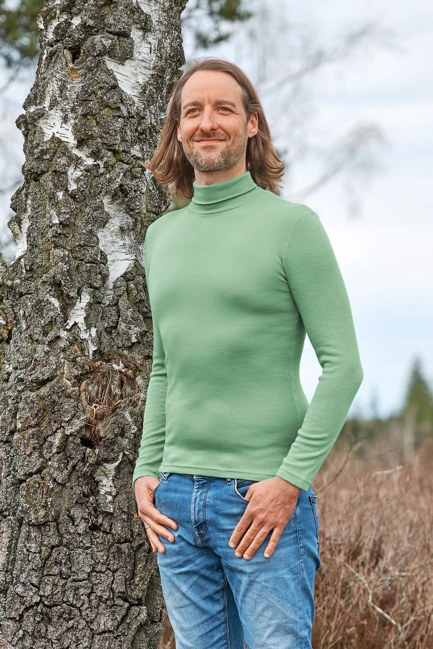 Mann mit Jeans steht neben Baum und trägt schilfgrünen Unterziehrolli aus bio Wolle (kbT) und Seide