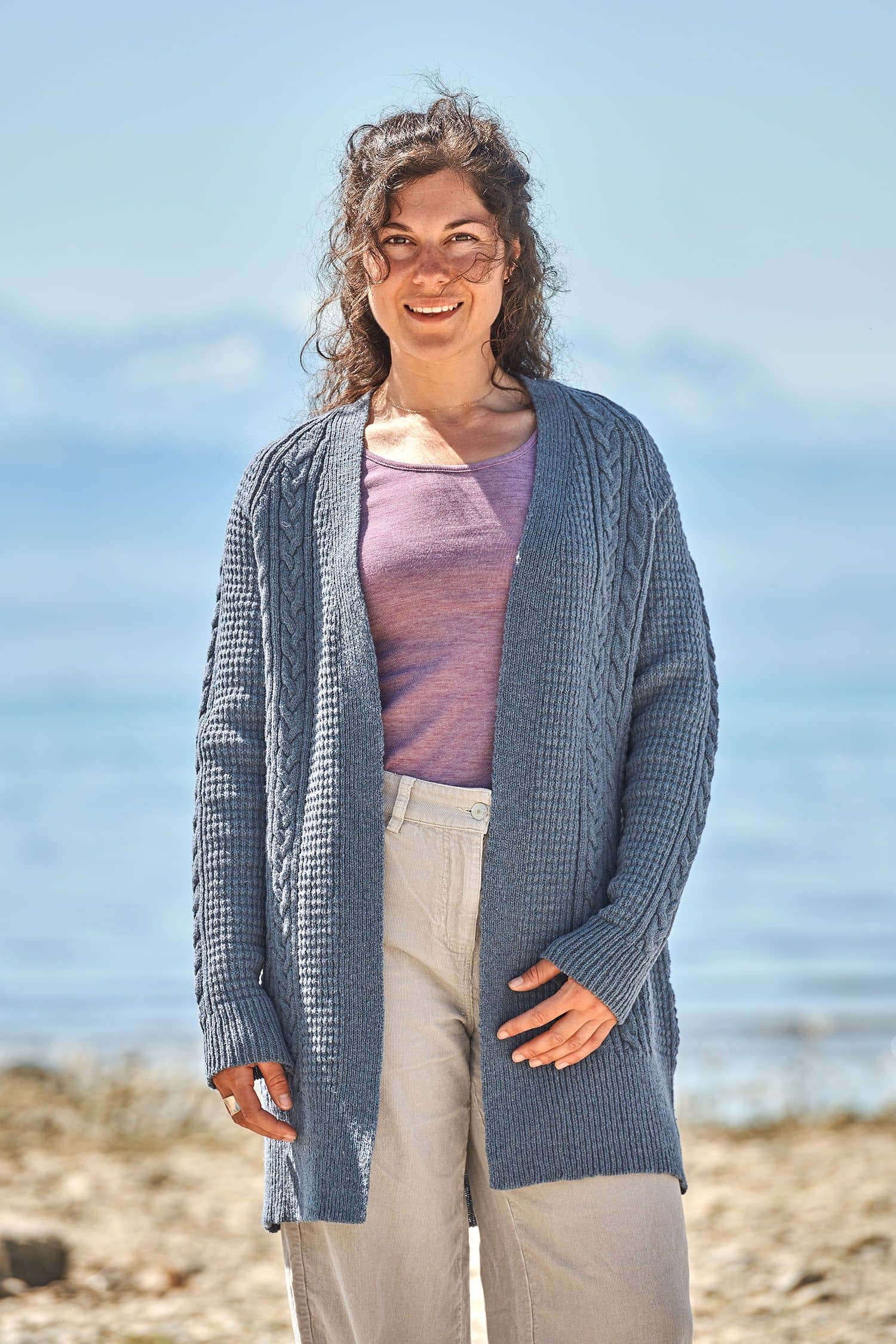 Frau trägt langen rauchblauen Cardigan mit Zopfmuster aus bio Wolle (kbT) vom Finkhof, eine beige Jeans und ein lila Shirt