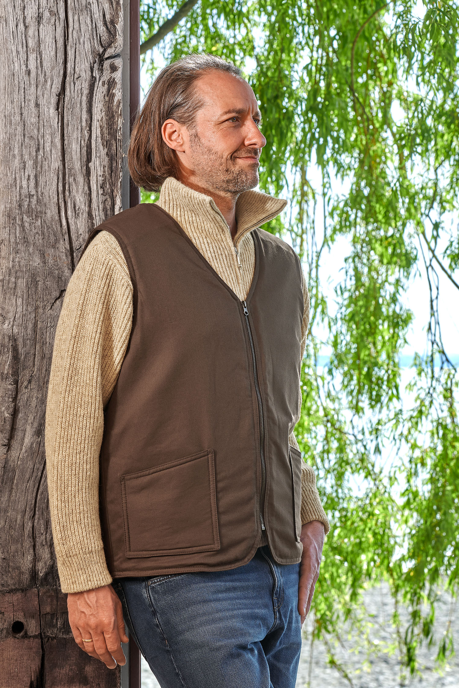 Mann lehnt sich an Baum und trägt einen Troyer und die braune Fellweste aus bio Baumwolle (kbA) und Fell vom Finkhof