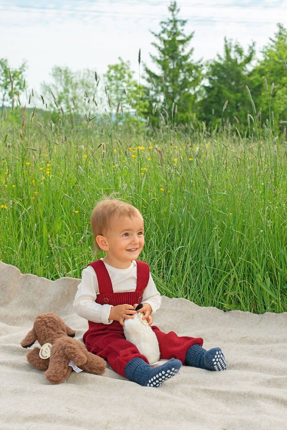 Kind sitzt mit Decke im Gras und trägt eine rote Hose und blaue Wollplüsch Stoppersocken aus bio Wolle (kbT)