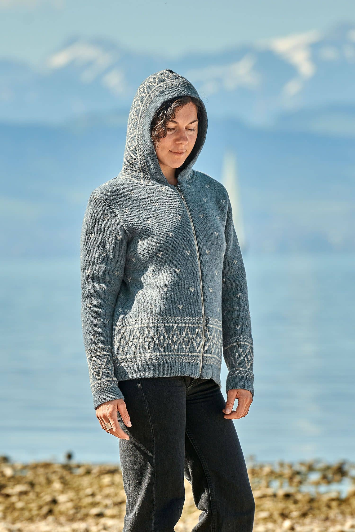 Frau vor Bergseekulisse trägt eine rauchblaue Norweger Kapuzenjacke aus bio Wolle (kbT) vom Finkhof und hat die Kapuze auf