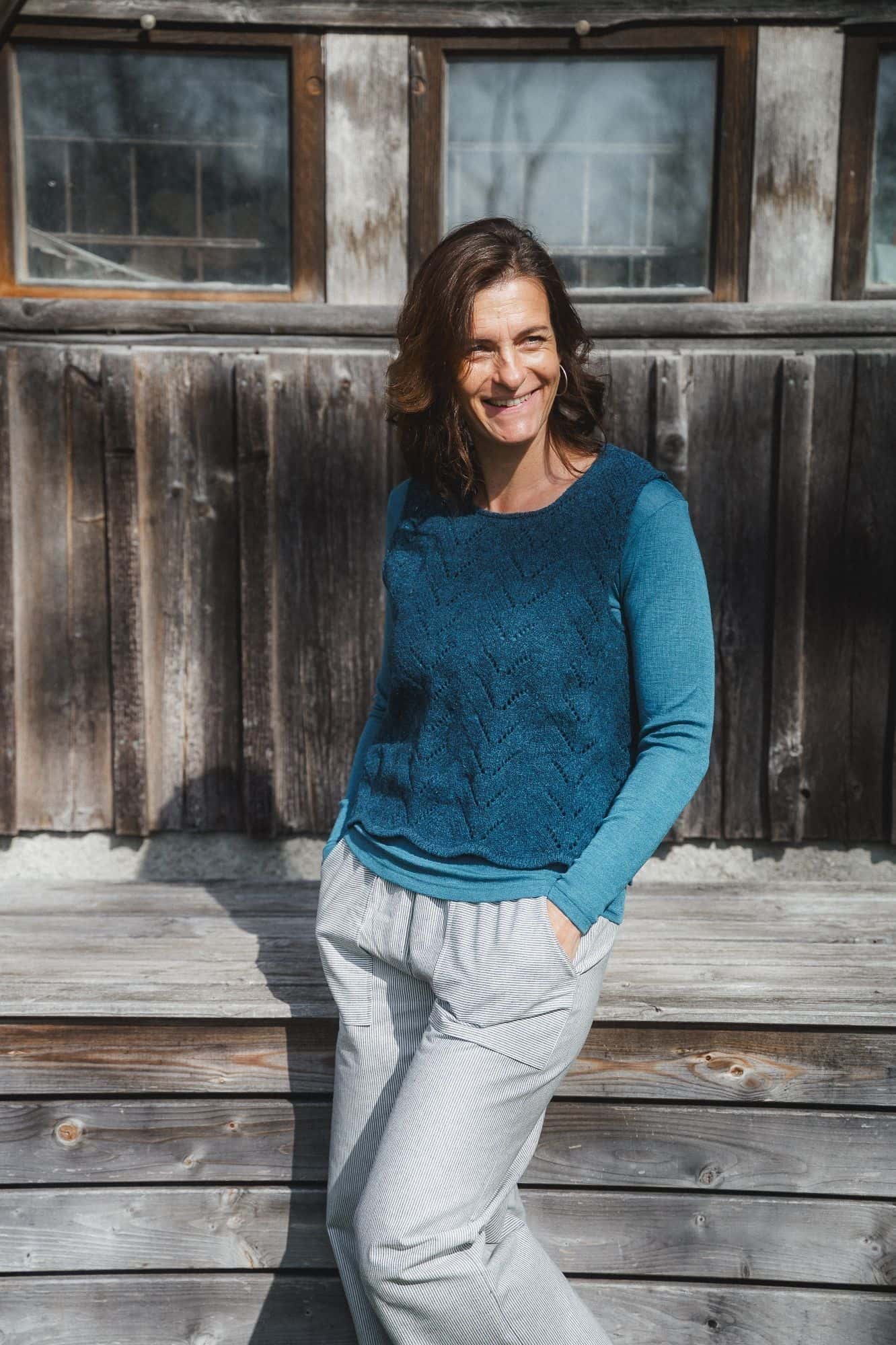 Finkhof-Ajour-Damen-Strickpullunder seeblau Gr. L Frau trägt blaues Langarmshirt und helle Baumwollhose mit blauem Strickpullunder aus Bio-Wolle vom Finkhof