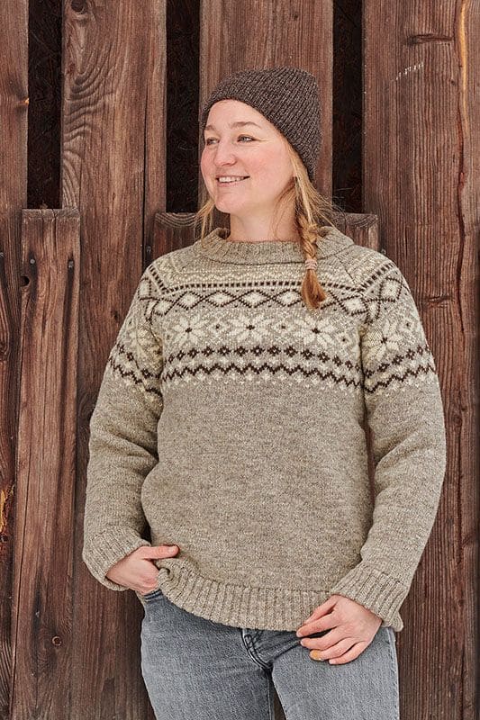 Frau steht vor Holzwand und trägt einen hellbraunen Norwegerpullover aus bio Wolle (kbT) vom Finkhof
