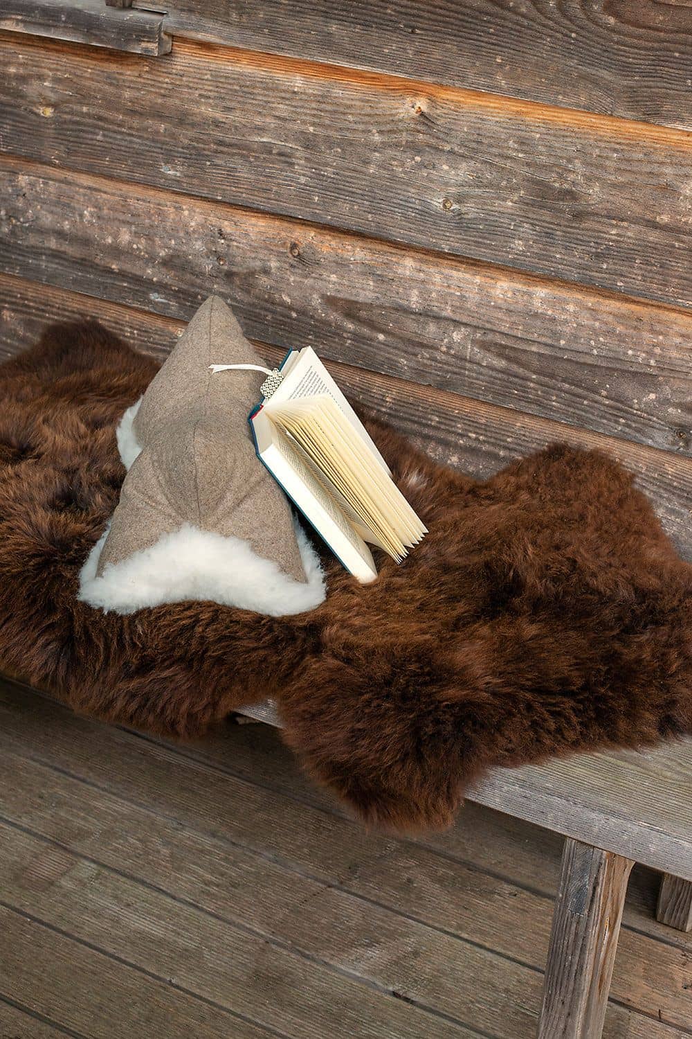 Leseknochen auf Holzbank liegt braunes Fell mit Buch und Finkhof Leseknochen aus pflanzlich gegerbtem Schaffell, Loden und Rohwolle (kbT)
