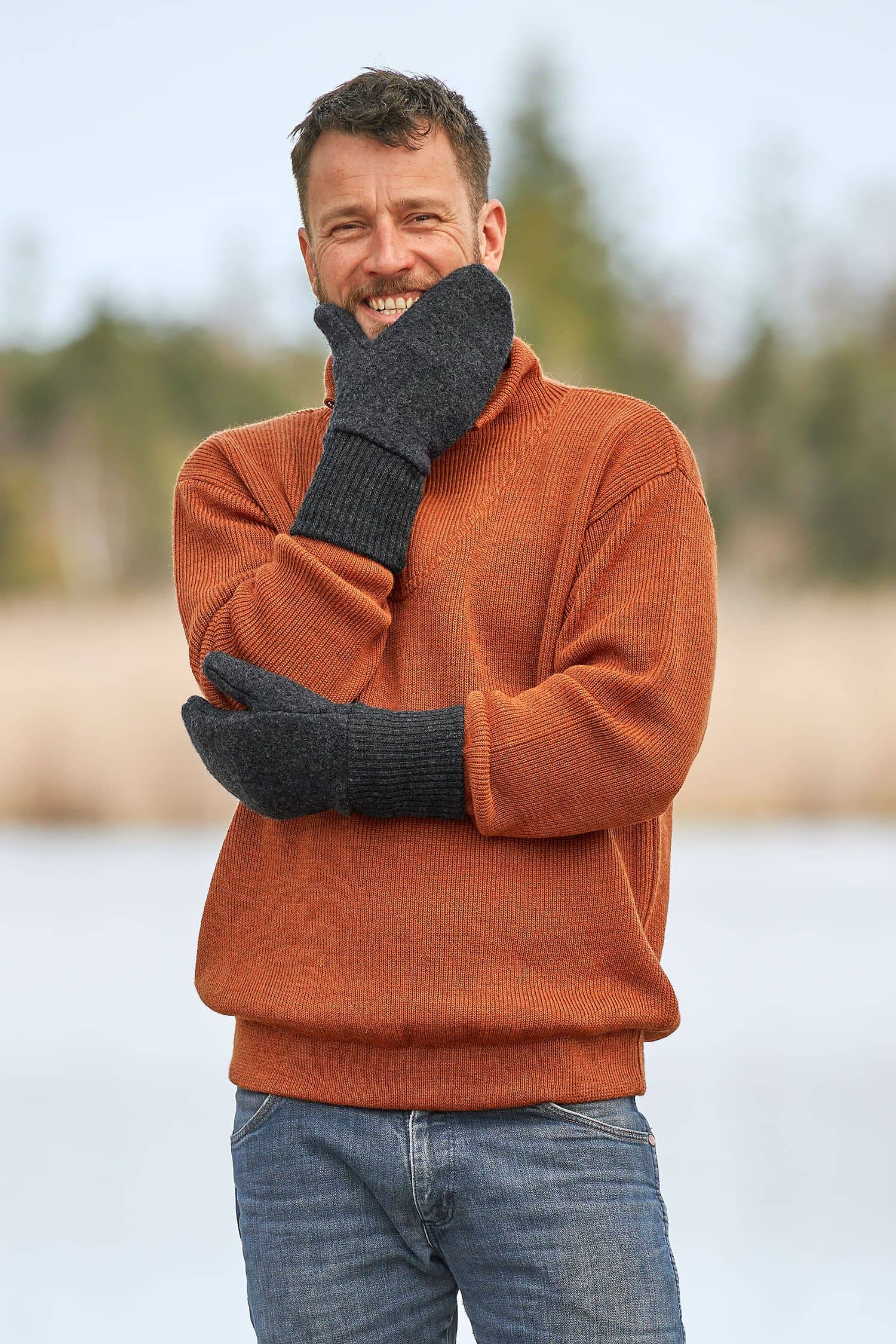 Mann mit orangem Strickpulli lacht und trägt dunkelgraue Walkfäustlinge vom Finkhof aus bio Wolle (kbT)
