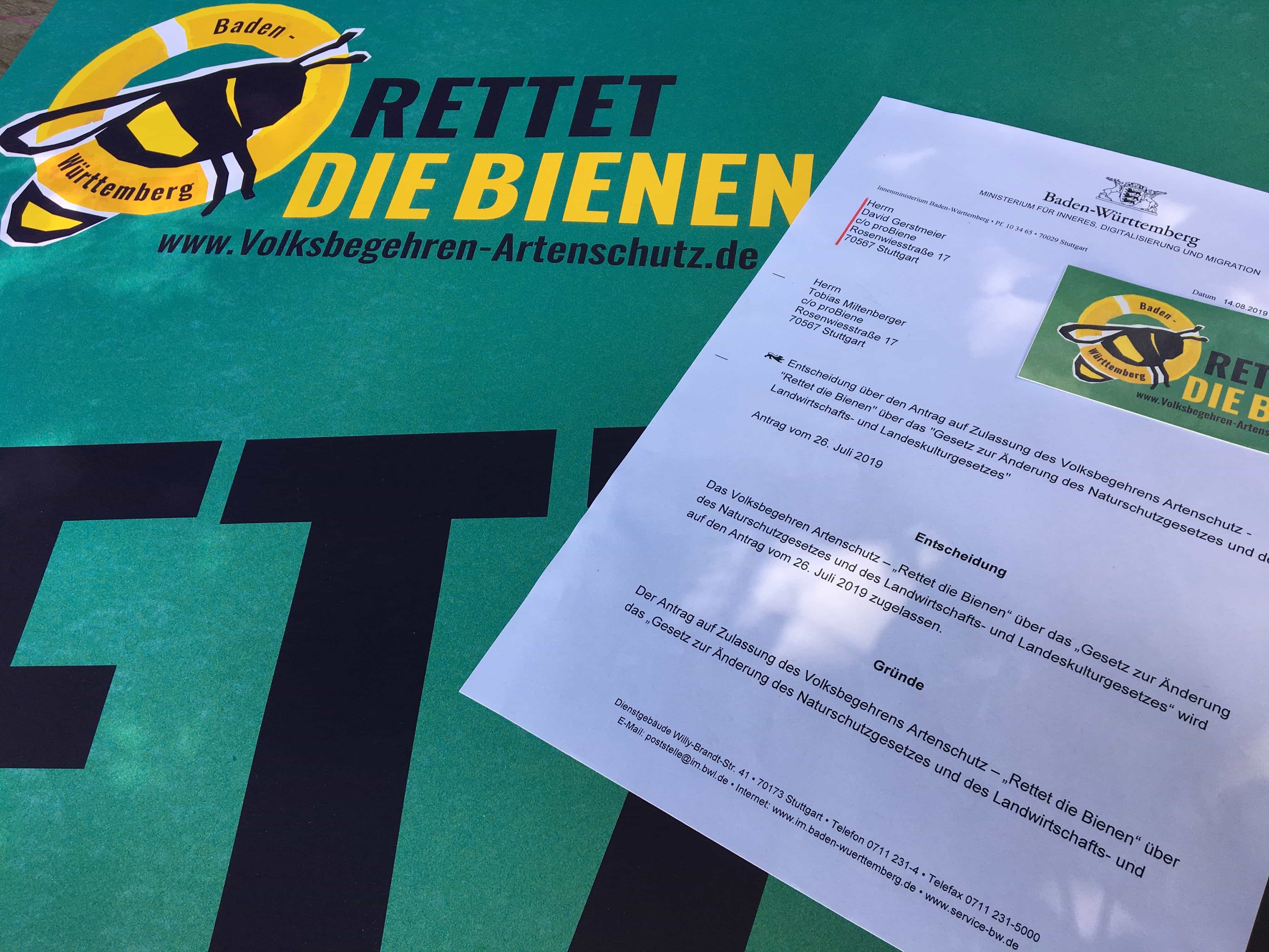 Entscheidungsdokument zum Antrag auf Volksbegehren für den Artenschutz liegt auf einem türkisen Rettet die Bienen Plakat
