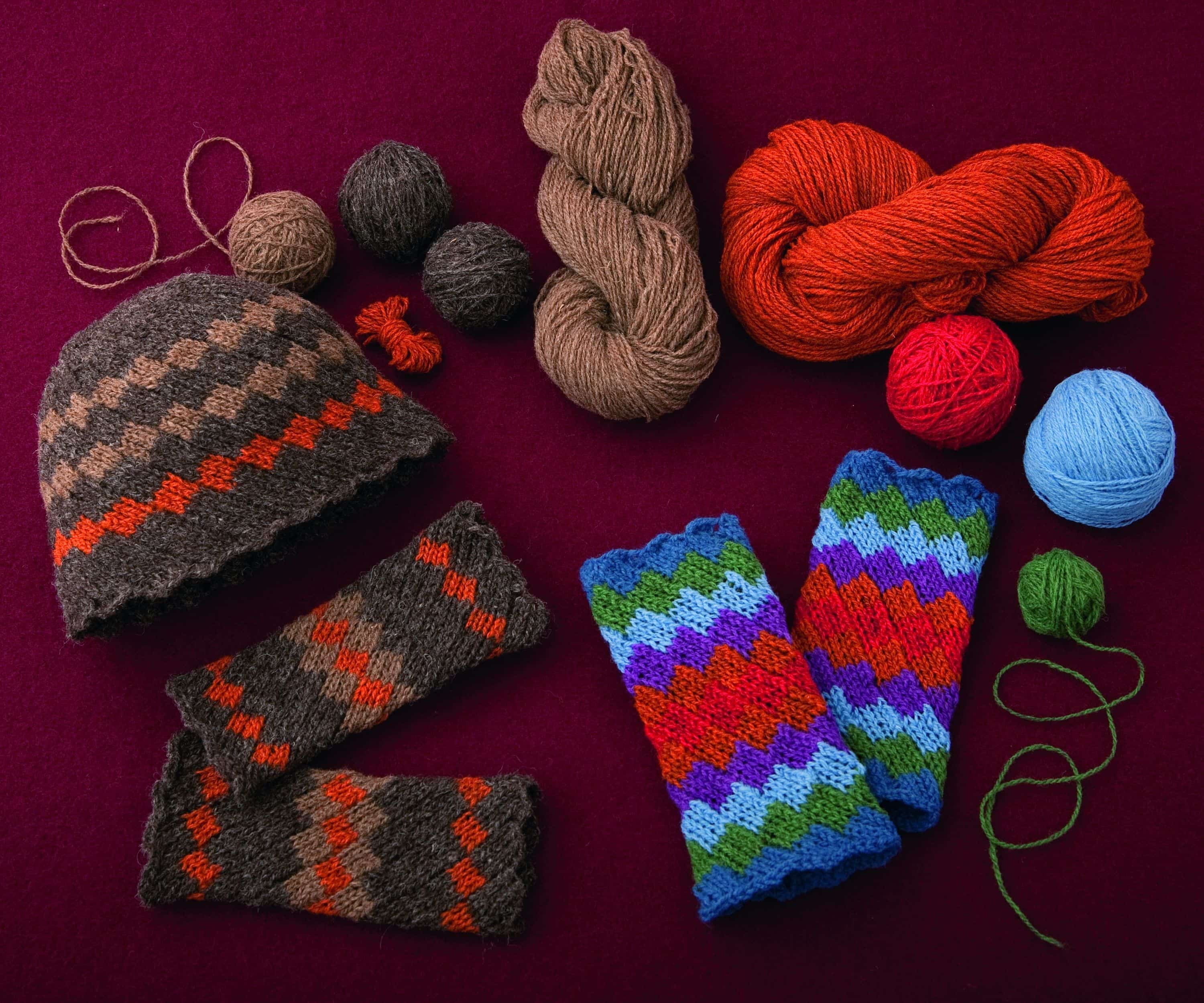 Mütze und gemusterte Stulpen liegen mit Wollknäulen auf rotem Untergrund
