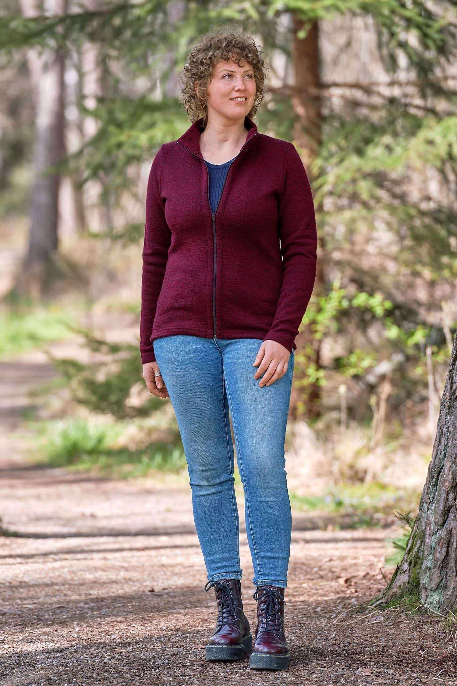Frau steht im Wald und trägt Jeans, blaues Shirt und beerefarbene gewalkte Jacke