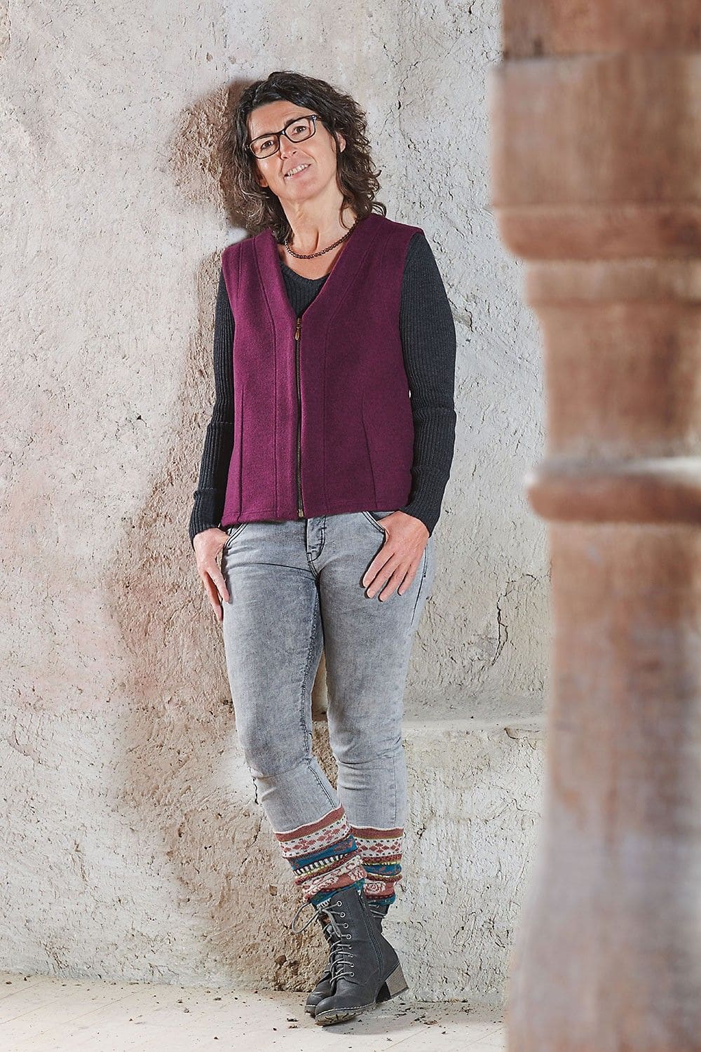 Frau lehnt an Wand und trägt graue Kleidung, brombeerfarbene Weste vom Finkhof und Beinstulpen