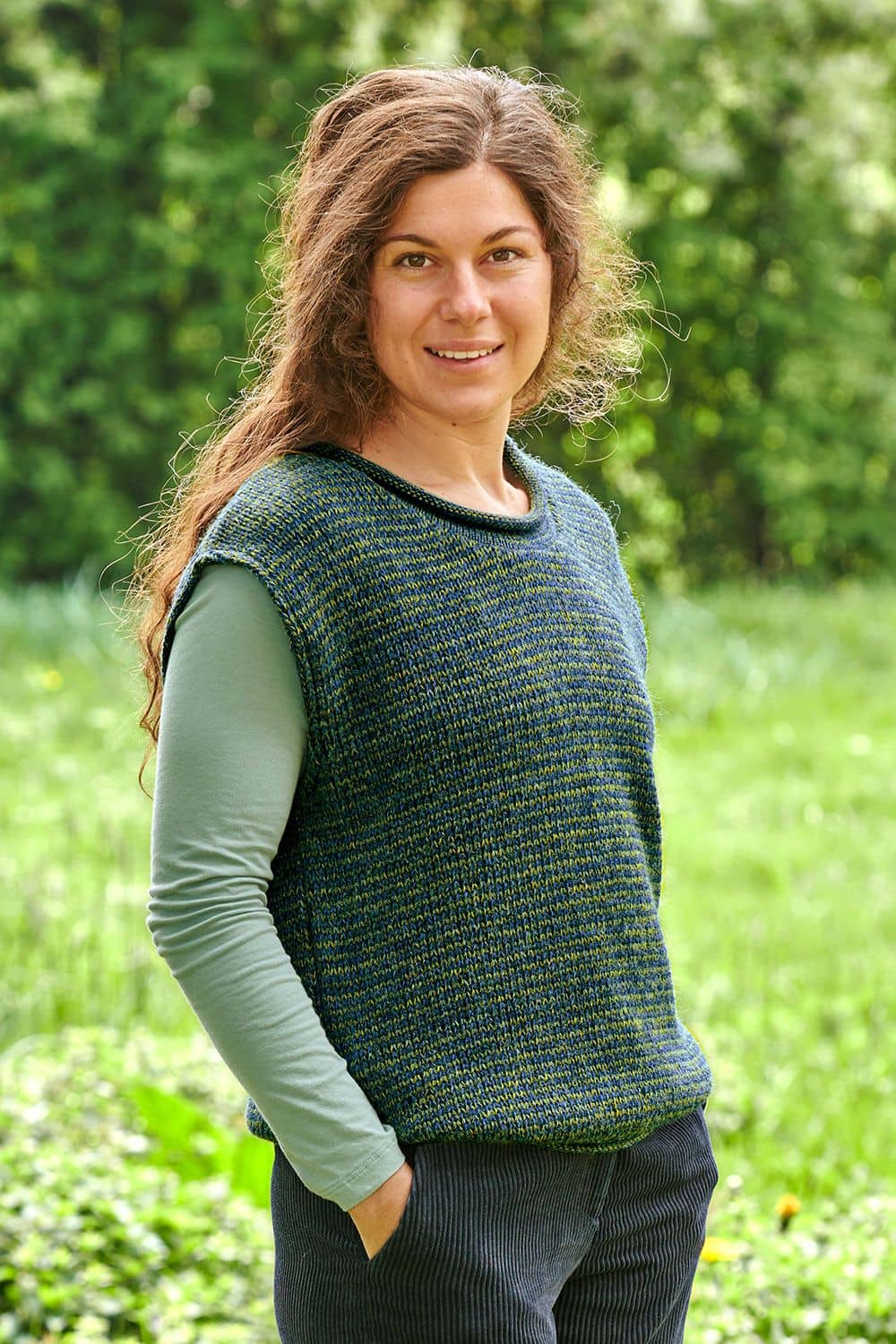 Frau auf Wiese mit hellgrünem, Shirt trägt blau/grünmelierten Pullunder aus bio Wolle (kbT)