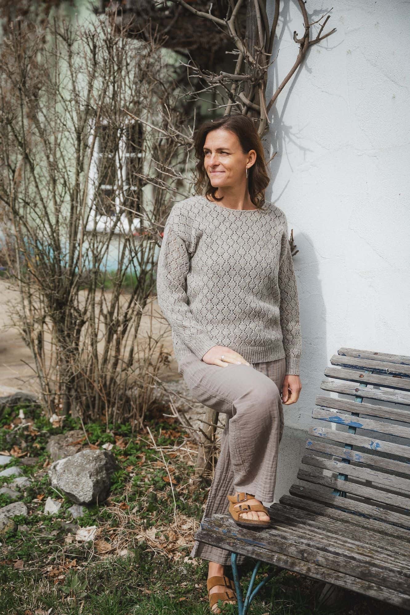 Finkhof-Ajour-Damen-Strickpullover ungefärbt Frau hat Fuß auf einer Bank stehen und trägt eine weite Hose und einen ungefärbten Strickpullover in grau mit Ajourmuster