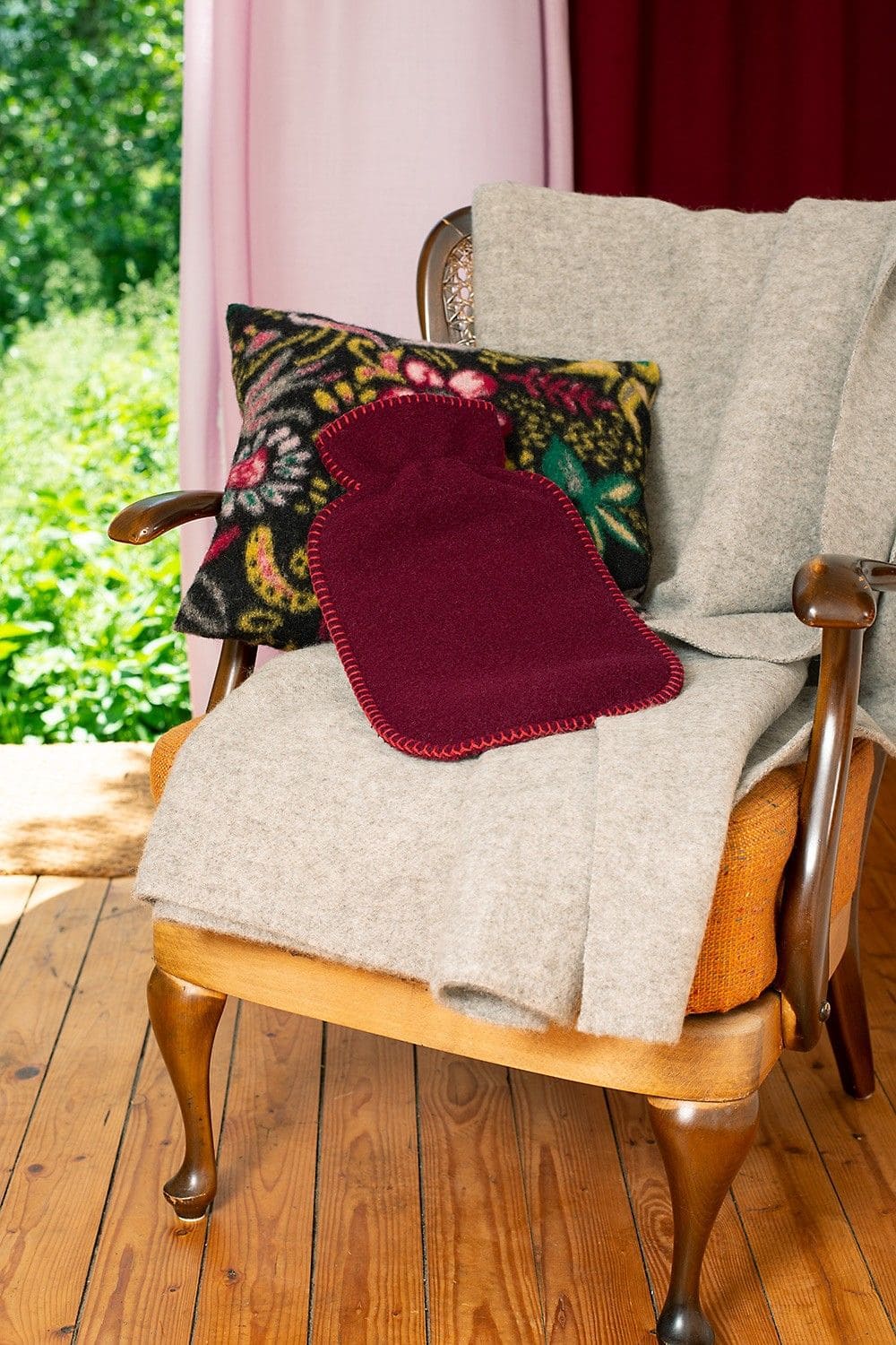 bordeauxroter Wärmflaschenbezug aus bio Wolle (kbT) vom Finkhof liegt auf Sessel mit Teppich, Kissen mit floralem Muster