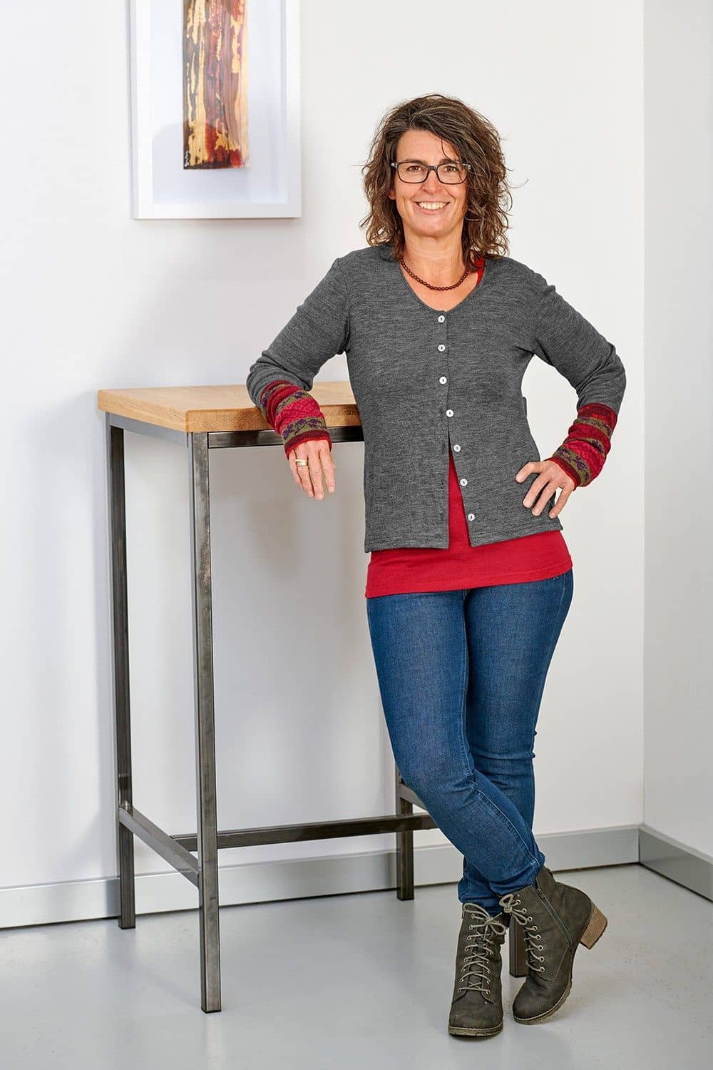 Frau lehnt sich an Tisch und trägt einen grauen Cardigan aus bio Wolle (kbT), rotem Shirt, gemusterten Stulpen und Jeans