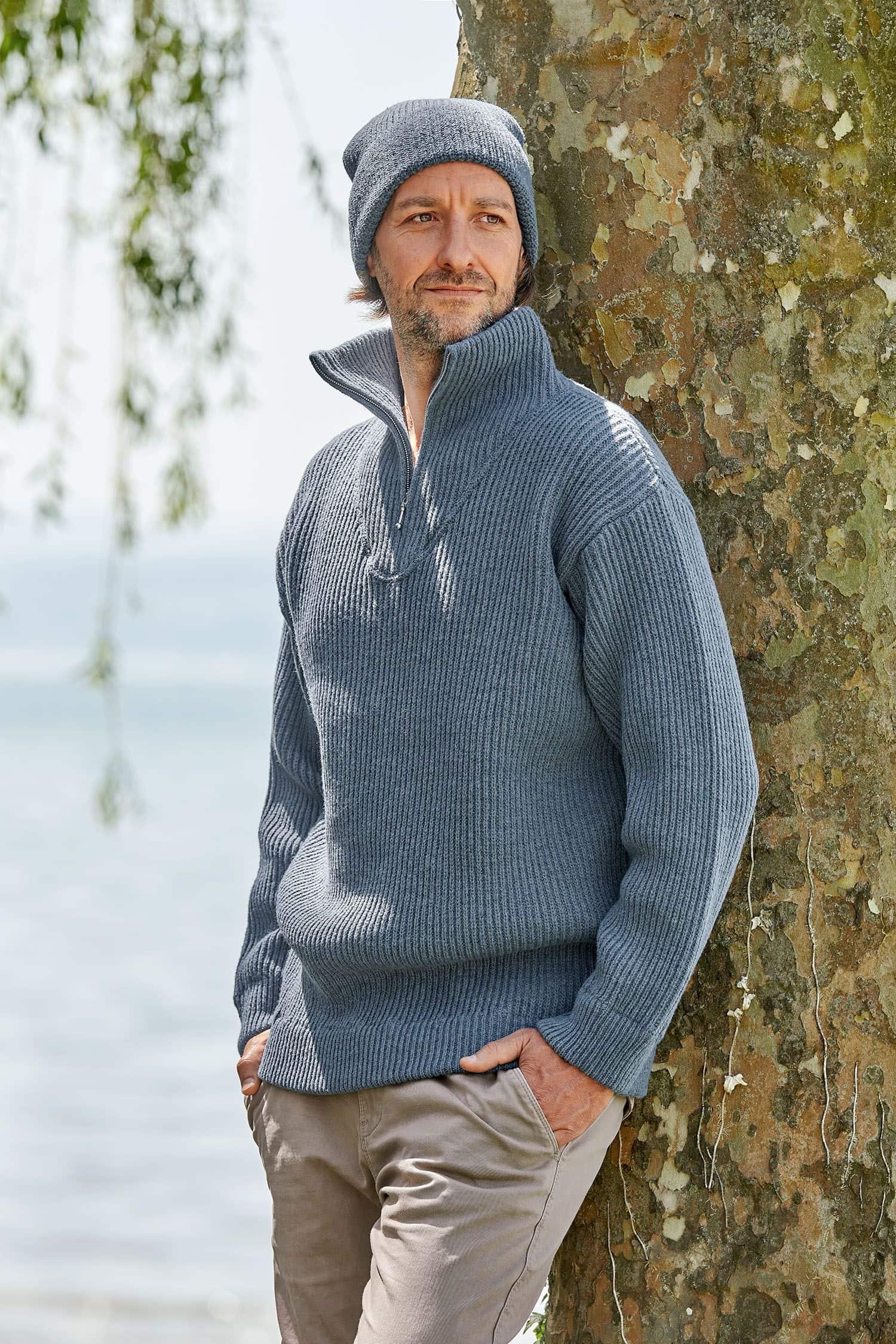 Mann lehnt sich an Baum und trägt eine rauchblaue Finkhof Mütze aus bio Wolle (kbT) und einen Troyer