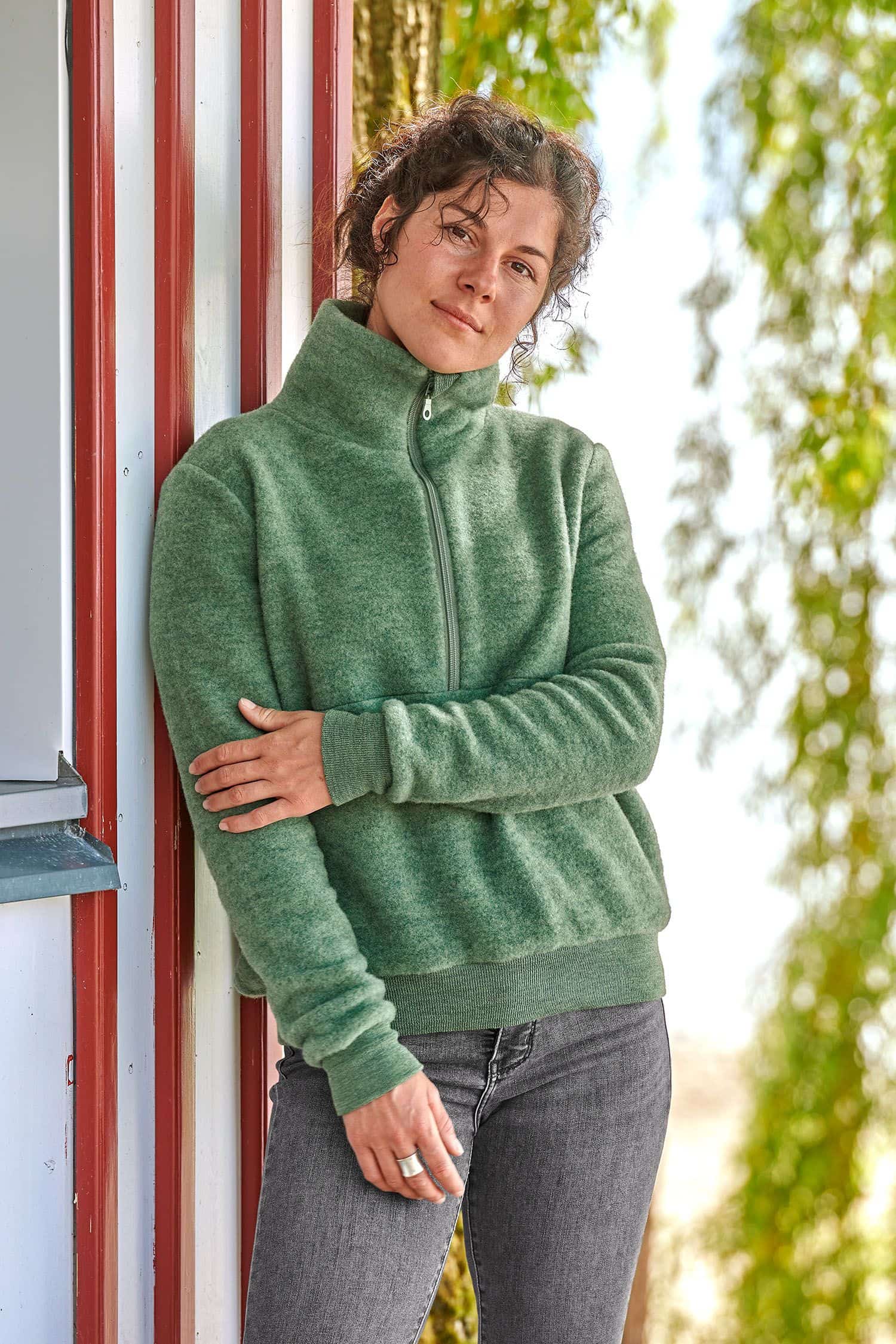 Frau lehnt an rot weißem Haus und trägt grünen Wollfleecepulli aus bio Wolle (kbT) vom Finkhof mit Reißverschluss