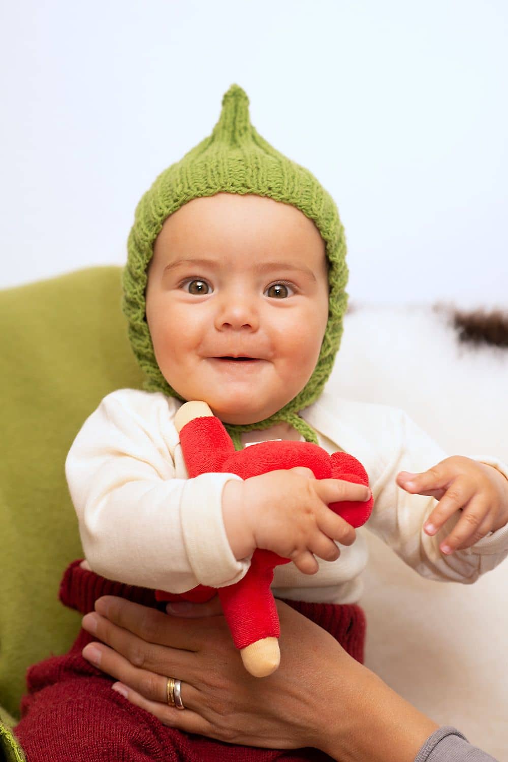 Kind trägt rote Wollhose, weißes Shirt und grüne gestrickte Zwergenmütze und sitzt dabei auf einem grünen Teppich