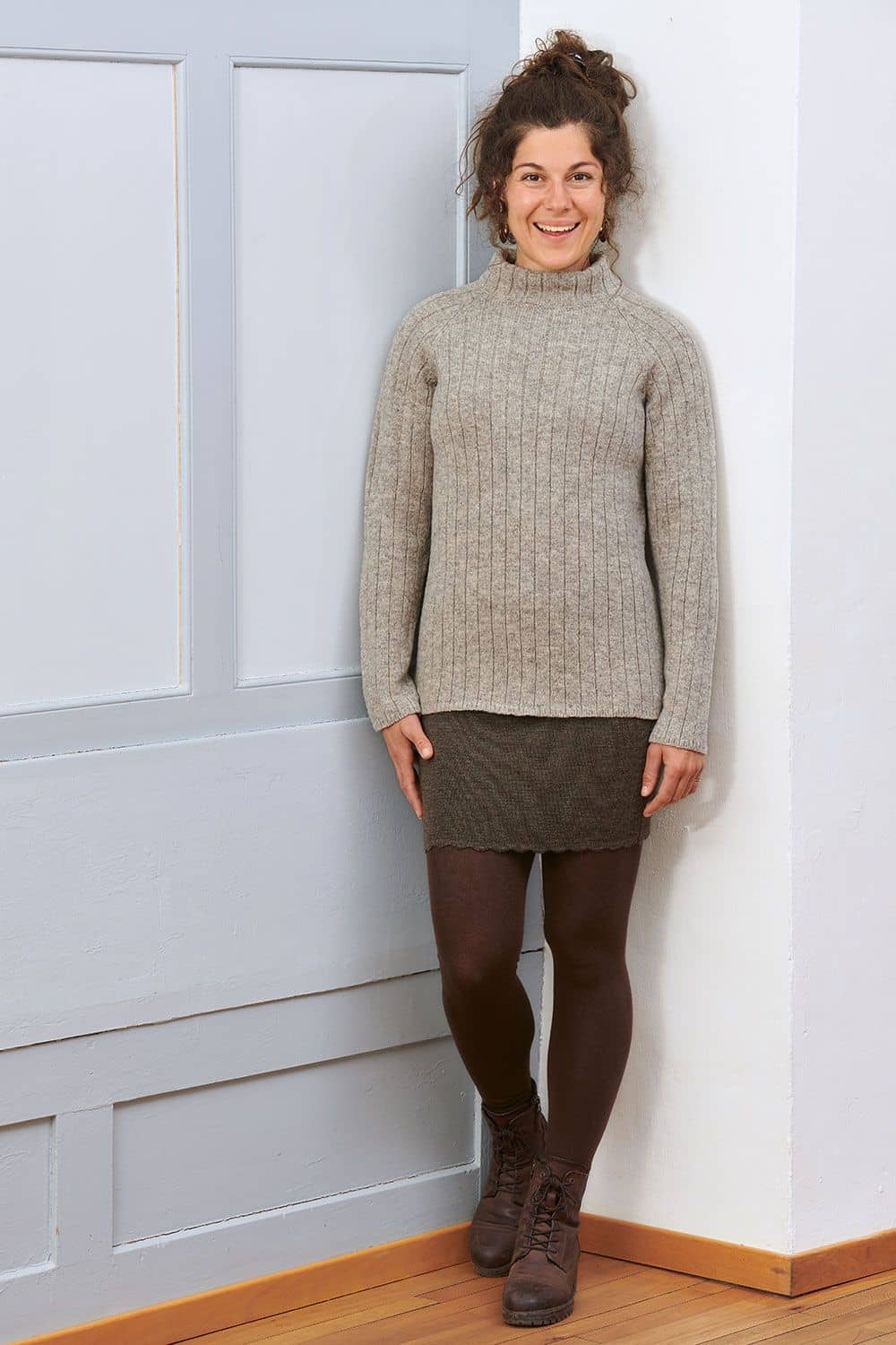 Frau steht lachend vor Wand und trägt grauen Reglanpullover vom Finkhof aus bio Wolle (kbT), braunen Rock und Stiefel