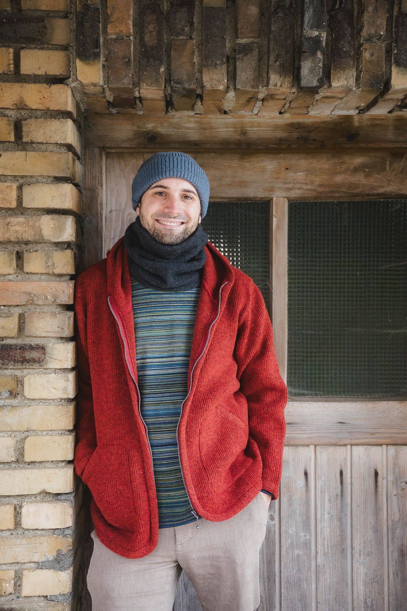 Mann mit Streifenshirt, Schal und Mütze trägt eine rote Wollwalkjacke mit Kapuze aus bio Wolle (kbT) vom FInkhof