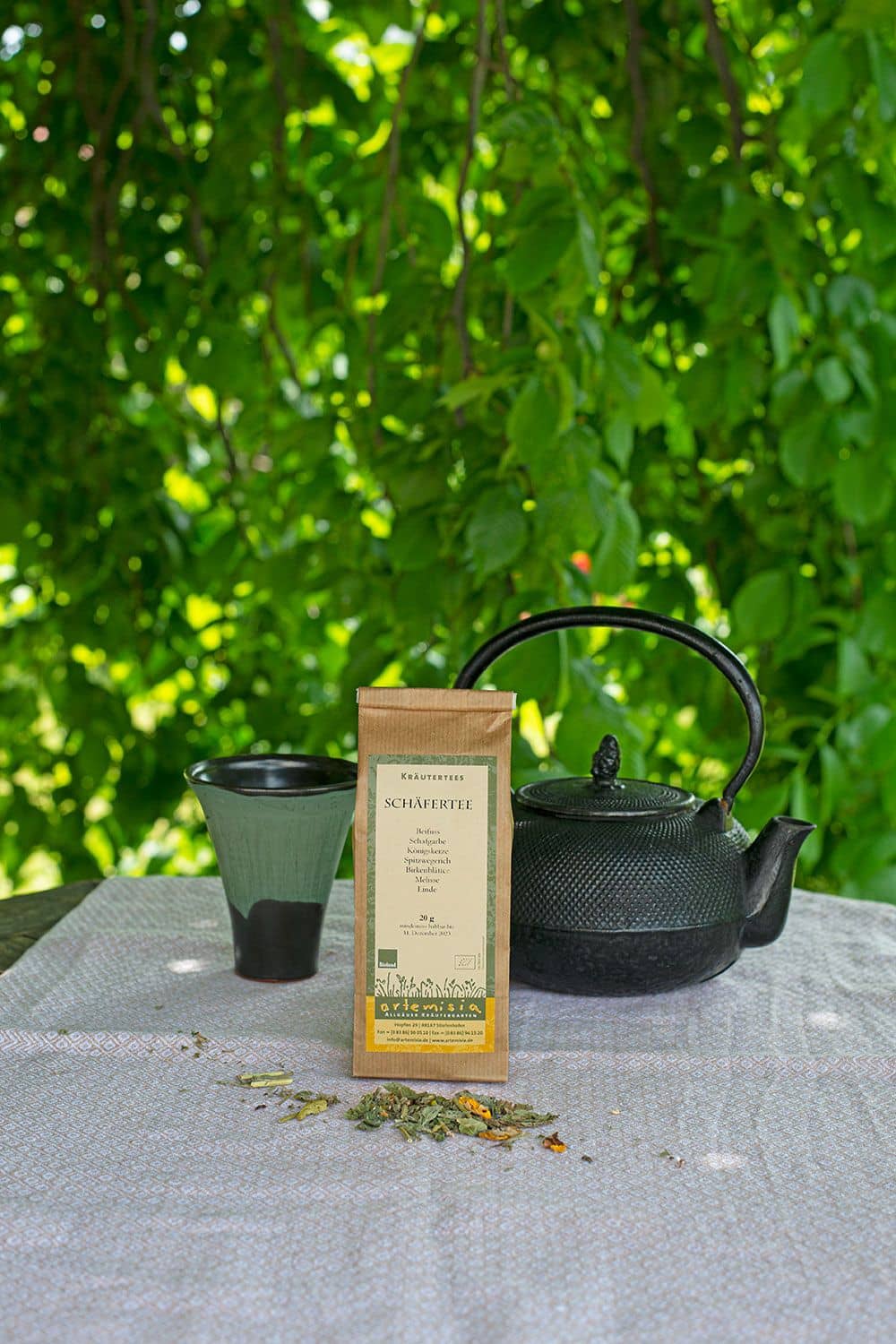 Schäfertee Schäfertee im 20g Pack auf Stimmungsbild unter Baum mit Teekanne, Becher du Kräutern auf weißer Tischdecke