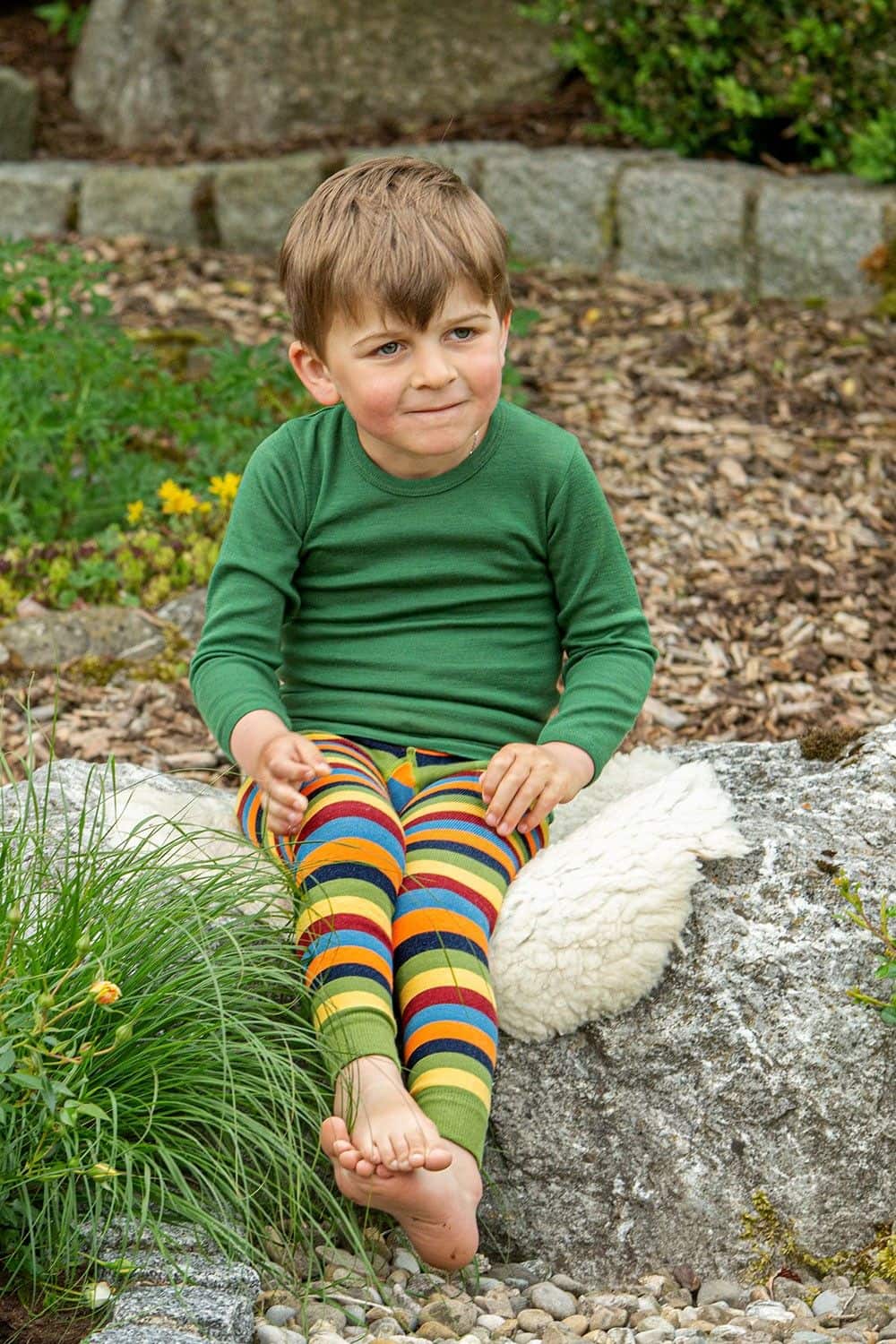 Kind mit grünem Shirt sitzt auf Stein und trägt bunt gestreifte Leggings aus bio Baumwolle (kbA) und Elasthan