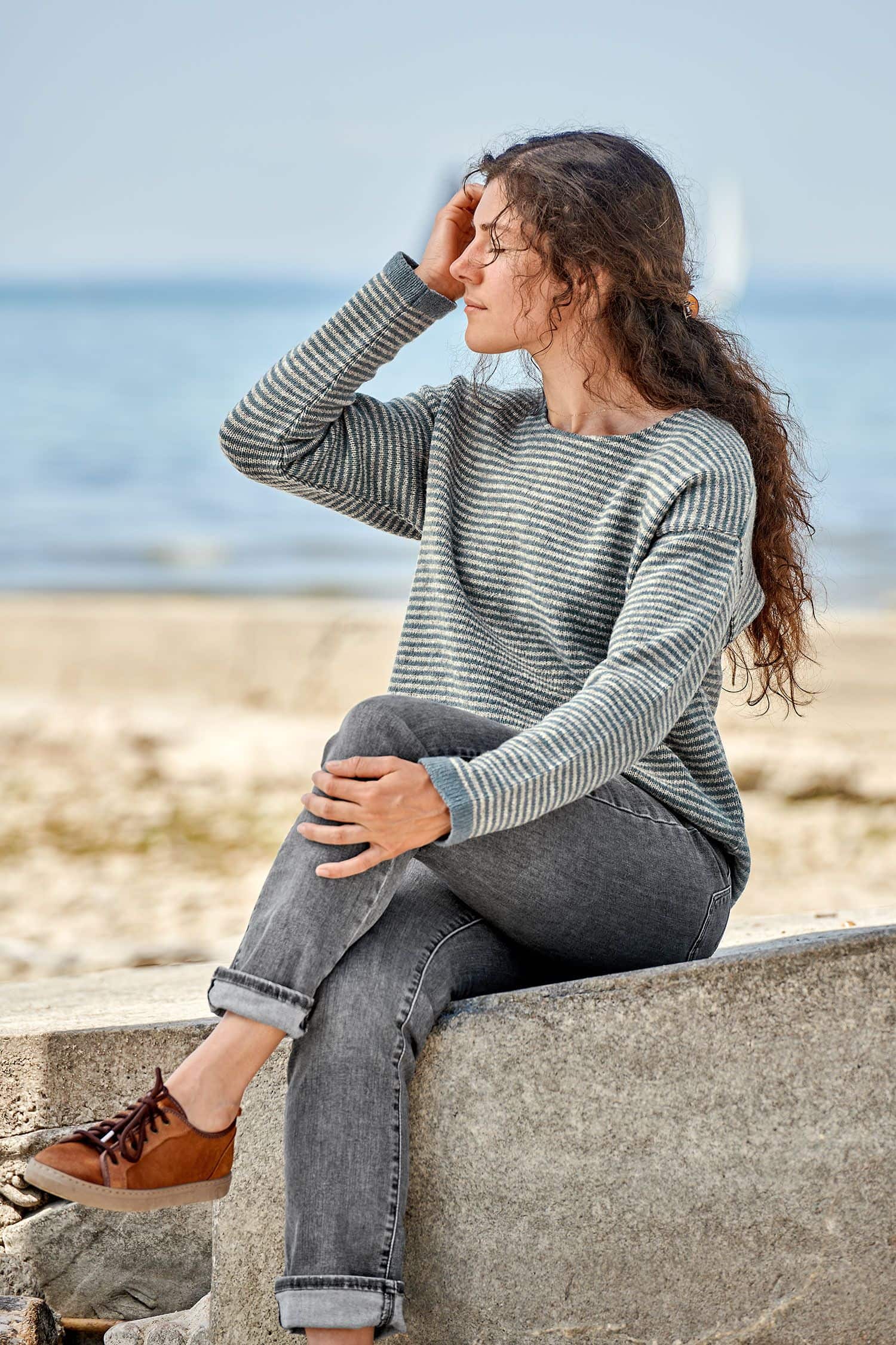 Frau sitzt auf Stein und trägt einen grau blauen Ringelpulli vom Finkhof aus bio Merinowolle (kbT)