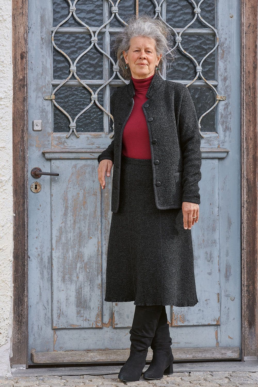 Frau trägt dunkelgrauen Wollrock und Woll Blazer mit rotem Shirt, Stiefeln und Strumpfhose und steht vor einer blauen Tür