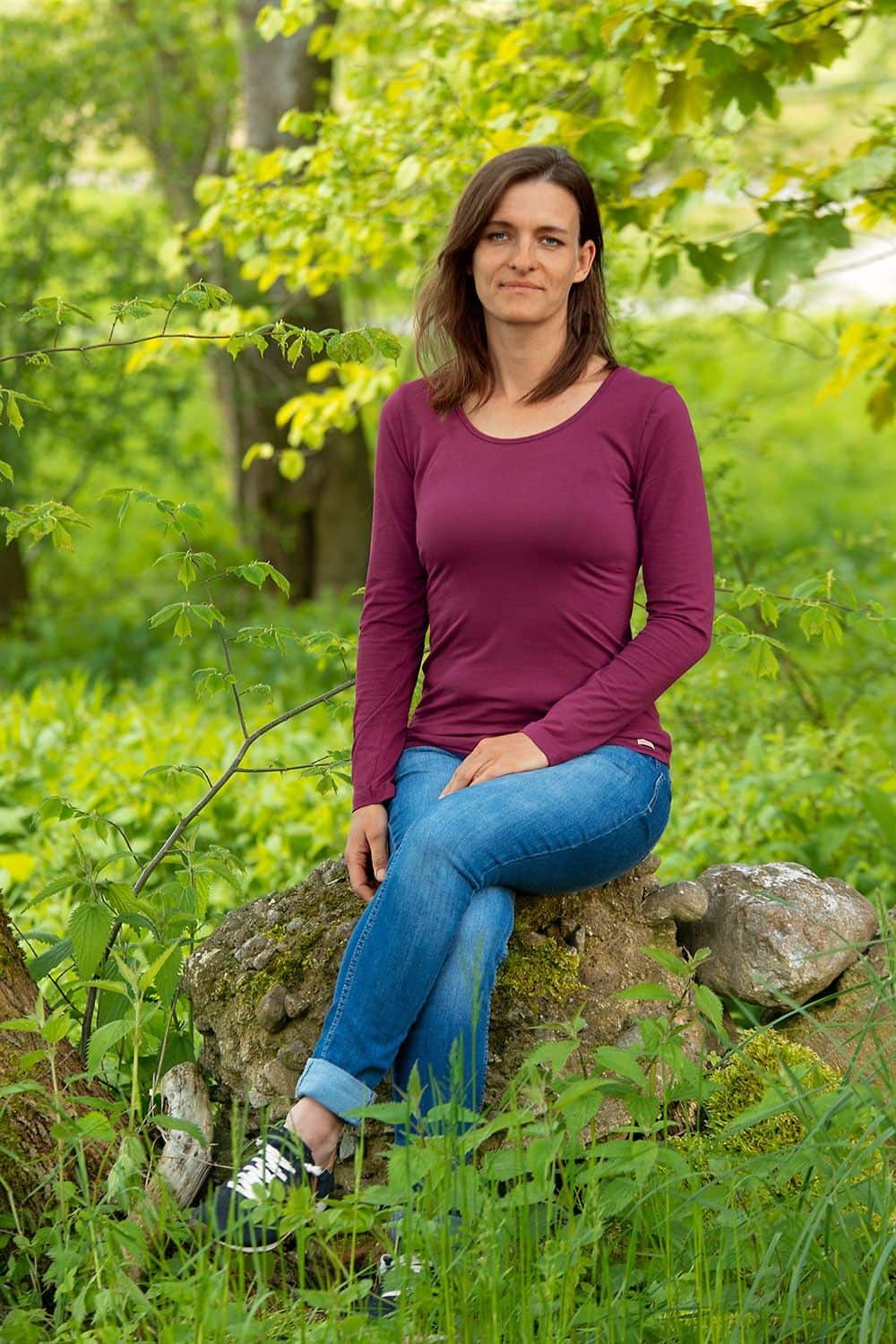 Frau sitzt auf Stein und trägt Jeans und brombeerfarbenes Langarmshirt