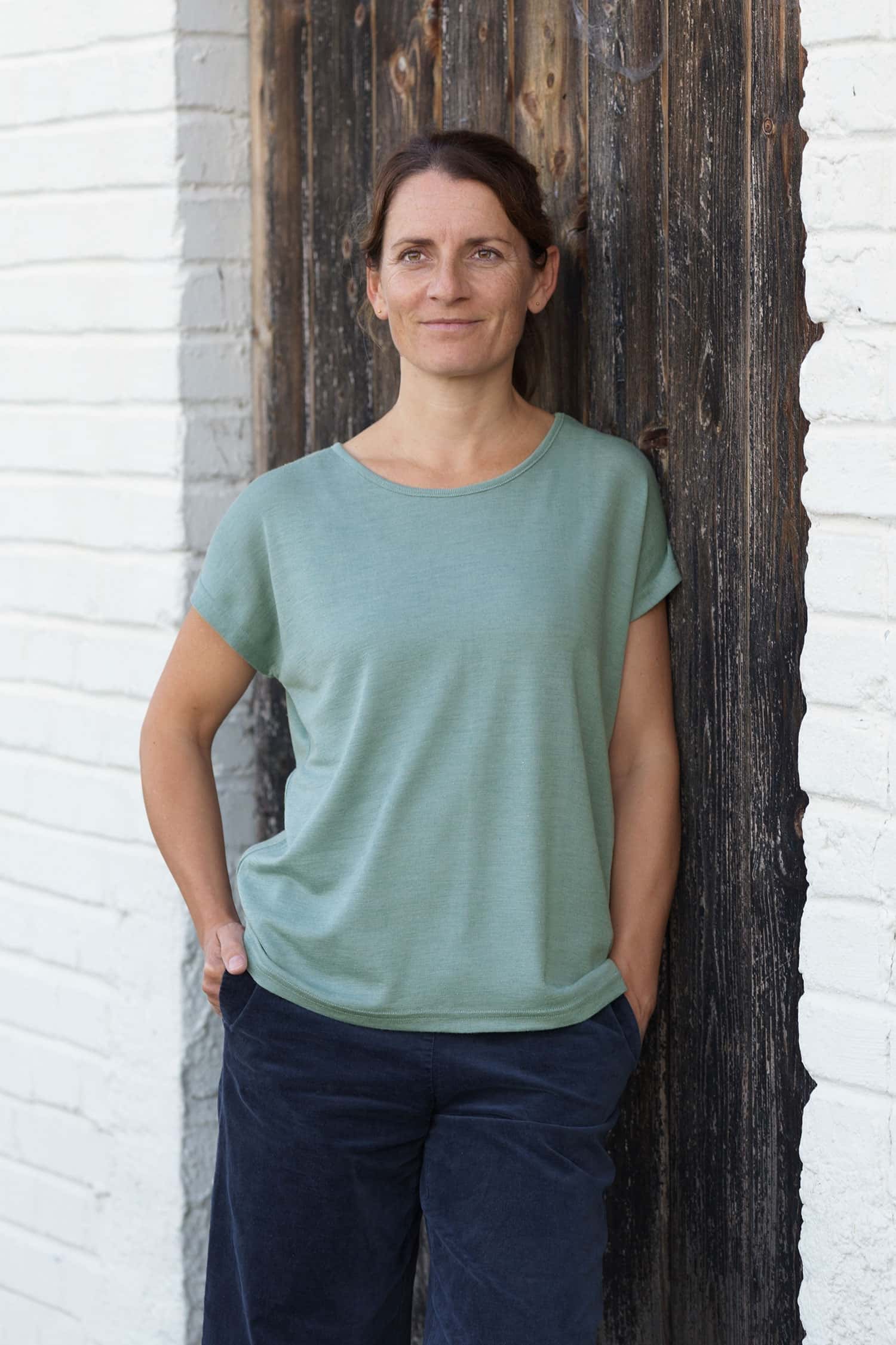 Frau lenht an Holztür und trägt eine dunkle Hose sowie ein pastellblaues Oversize Shirt aus Bio-Wolle und Seide