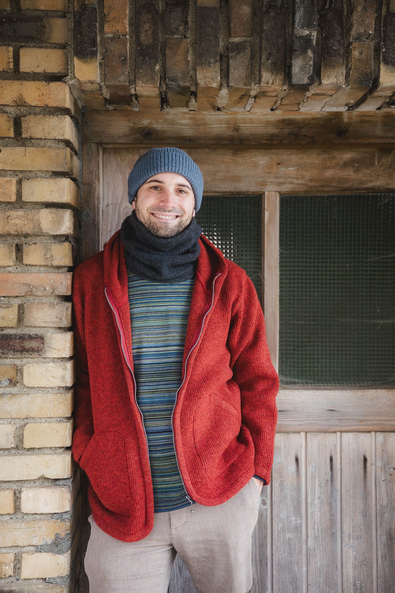 Mann mit Streifenshirt, Schal und Mütze trägt eine rote Wollwalkjacke mit Kapuze aus bio Wolle (kbT) vom FInkhof