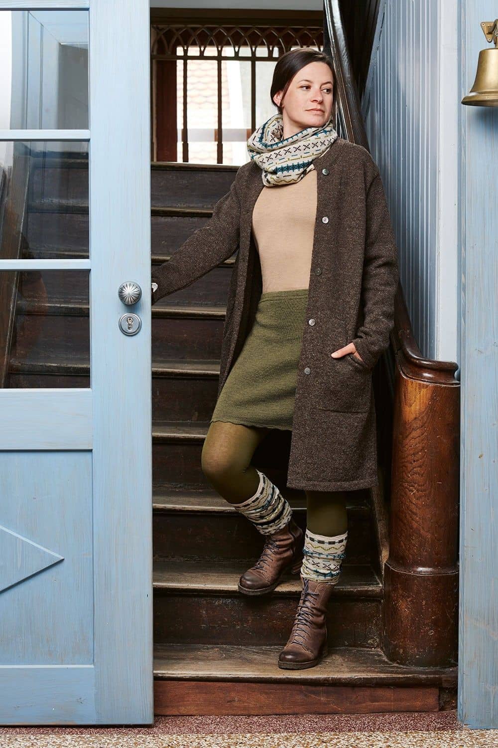 Frau auf Treppe trägt grünen Rock, Shirt, gemusterten Stulpen, Schal und braunen Finkhof Strickmantel aus bio Wolle (kbT)