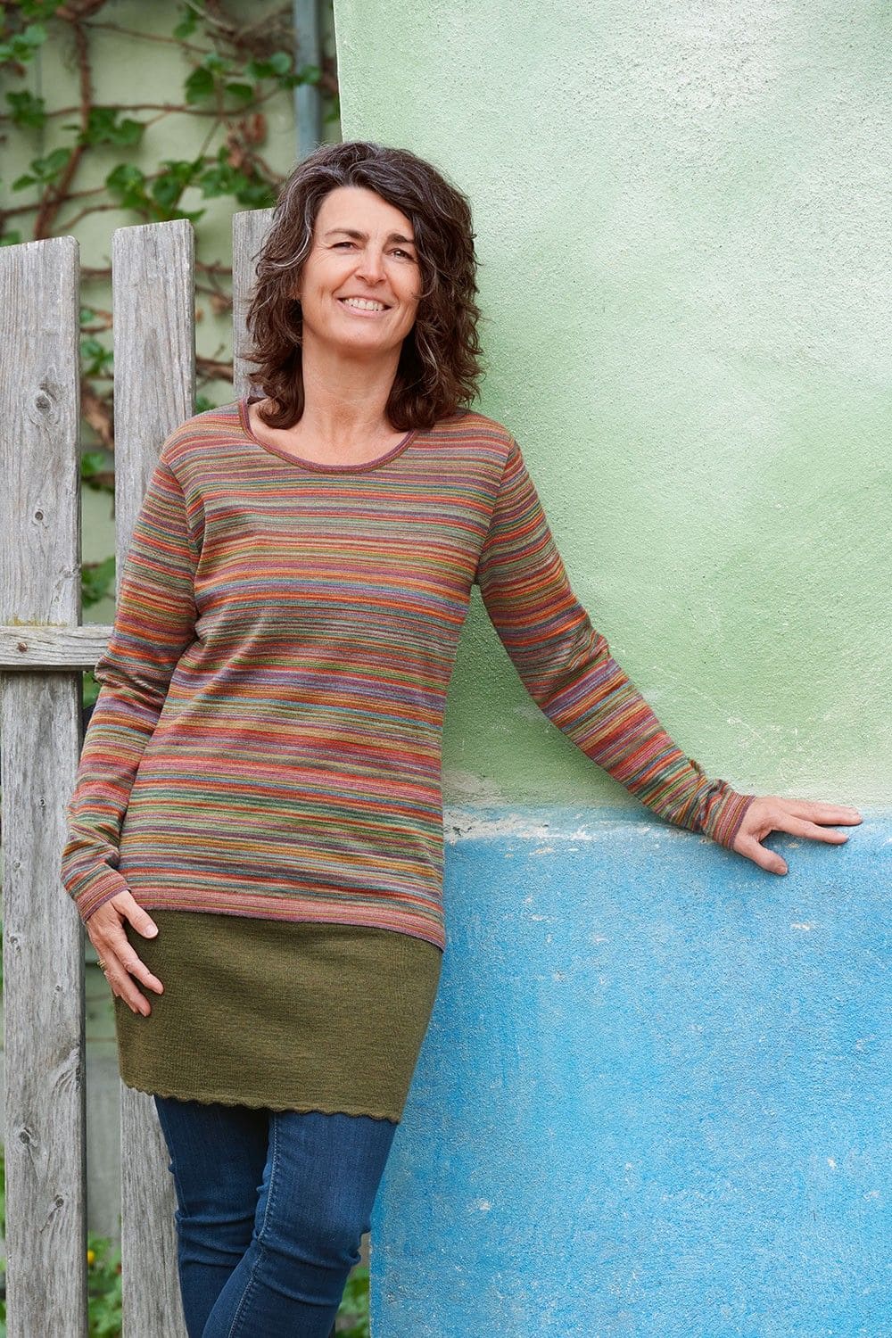 Frau mit pink/ buntem Ringelshirt aus bio Wolle (kbT) vom Finkhof und grünem Rock steht vor einer grünen Hauswand