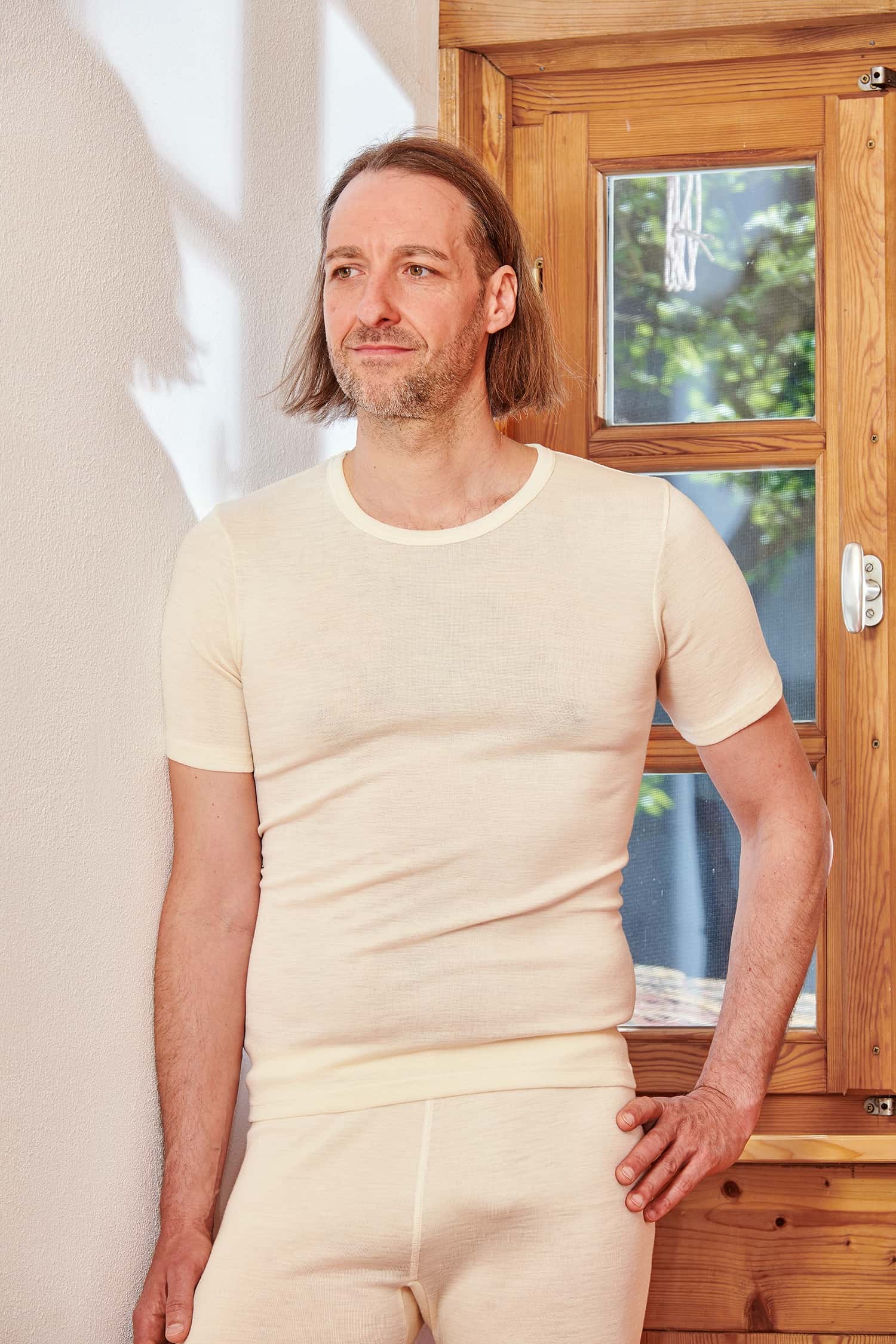 Mann lehnt an Tür und trägt ein naturweißes Kurzmarmshirt aus bio Wolle (kbT) und Seide und eine weiße lange Unterhose