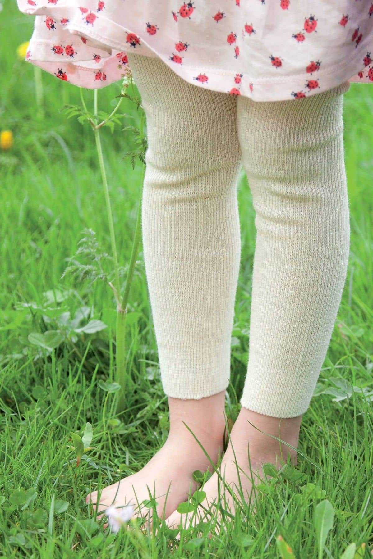 Kind steht auf einer Wiese und trägt eine naturweiße Woll-Leggings aus bio Wolle (kbT) und ein Kleid mit Marienkäfermuster