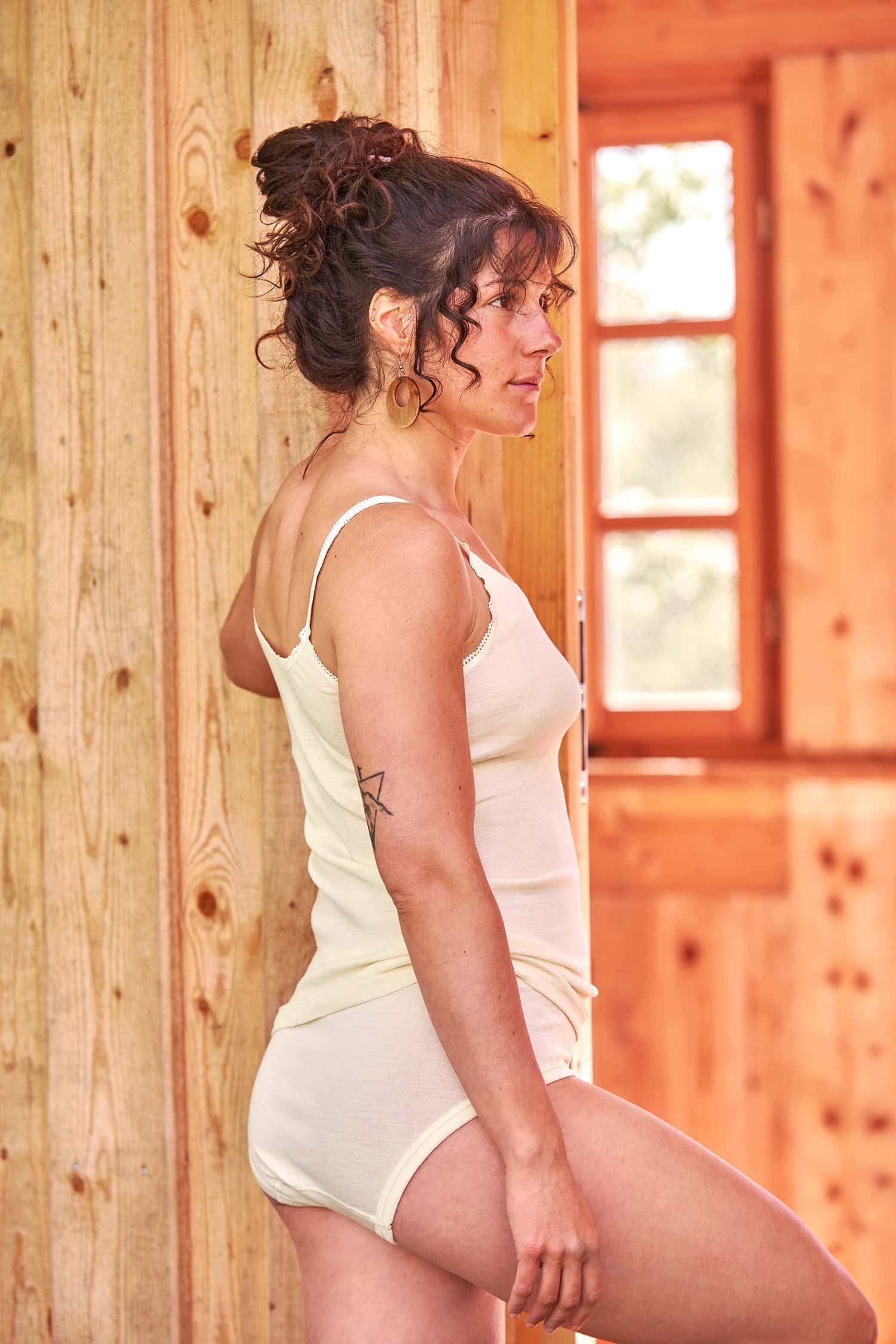 Seitenansicht von Frau vor Holzwand, die einen naturweißen Slip aus Bio-Wolle und Seide mit passendem Unterhemd trägt
