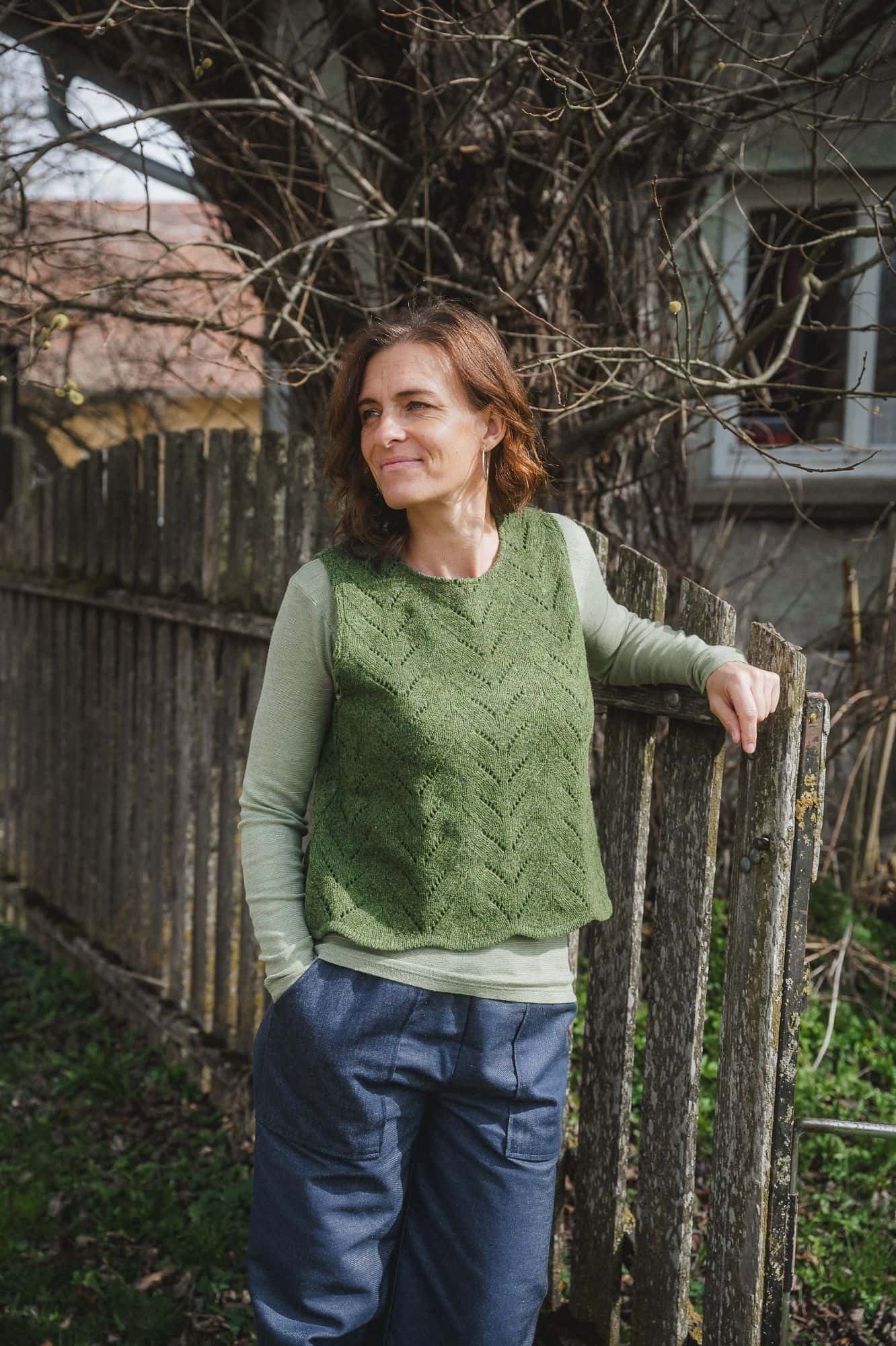 Finkhof-Ajour-Damen-Strickpullunder grün Gr. M Frau steht am Gartenzaun und trägt grünes Langarmhirt, blaue Hose und grünen Strickpullunder vom Finkhof