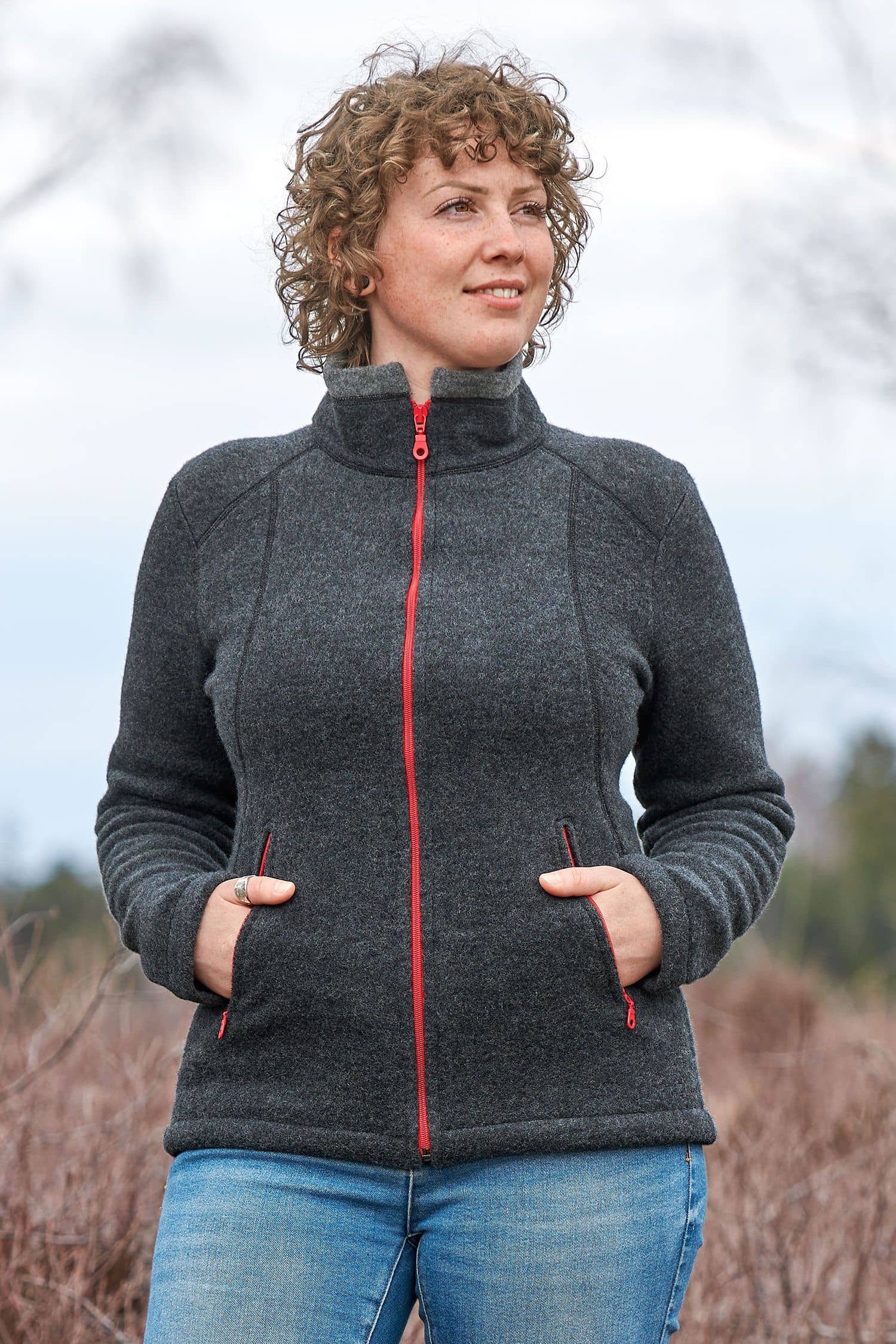 Frau mit Jeans trägt eine dunkelgraue Wollfleecejacke mit Stehkragen und roten Reißverschlüssen aus bio Wolle (kbT)