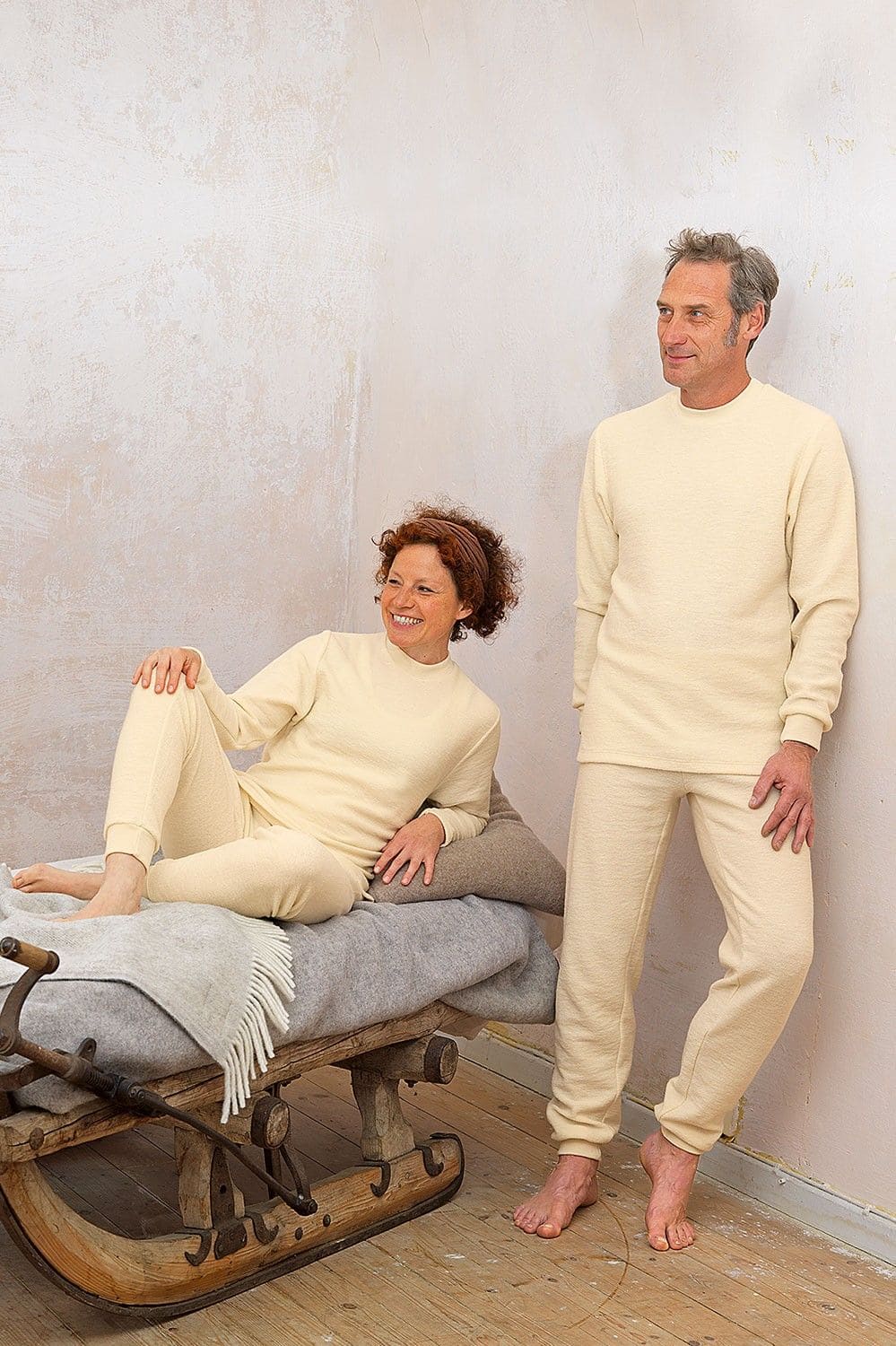 Mann lehnt sich an Wand,Frau liegt auf Holzschlitten beide tragen naturweiße Wollfrotte-Schlafanzüge aus bio Wolle (kbT)