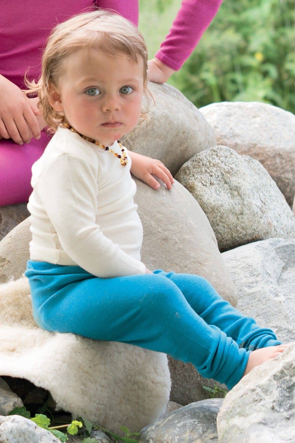 Kind sitzt auf Stein und trägt eine türkise Hose und ein naturweißes Langarmshirt aus bio Wolle (kbT) und Seide