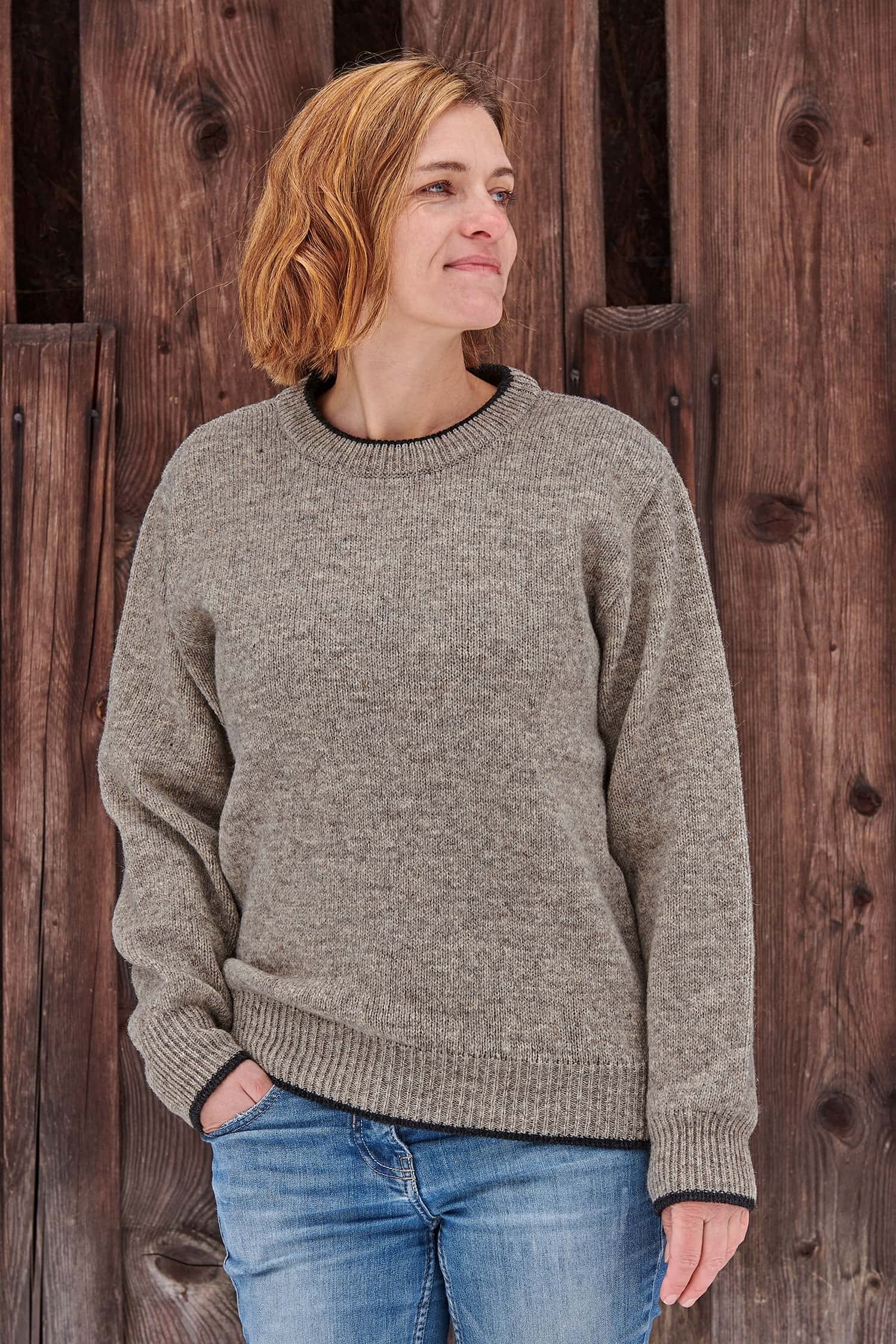 Frau vor Holzwand trägt grauen Finkhof Pulli aus bio Wolle (kbT) mit Rundhalsausschnitt und blauer Jeans