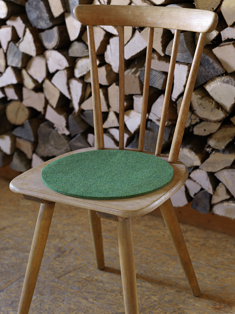 grüne runde Finkhof Sitzauflage aus bio Wolle (kbT) auf einem Stuhl vor einer Holzwand