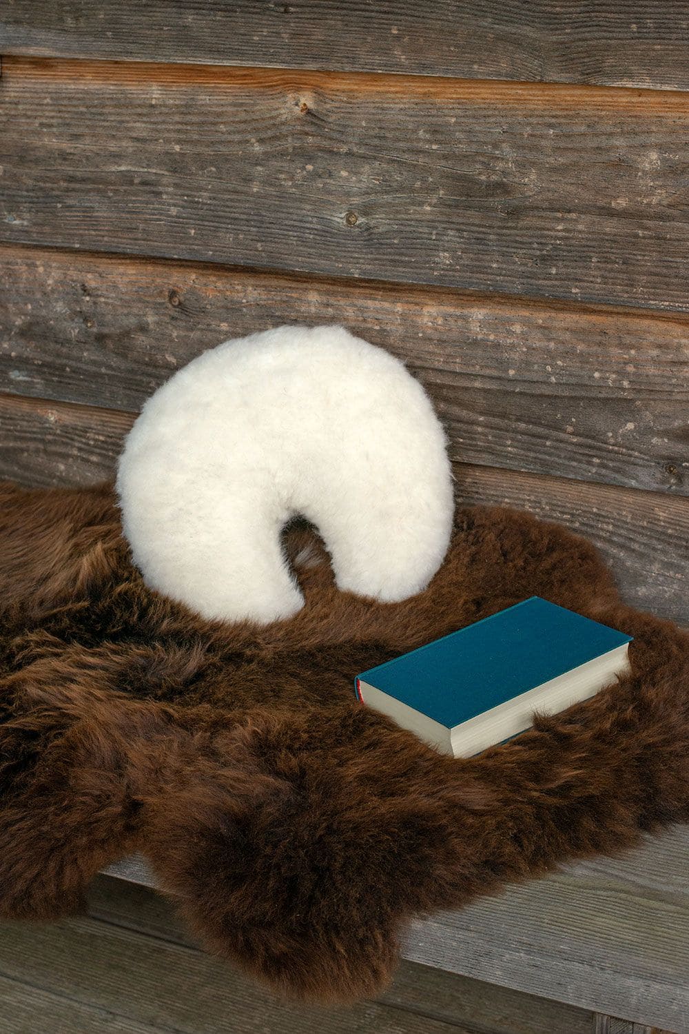 weißes Finkhof Stützkissen in Mondform aus Schaffell liegt mit Buch und Brille auf braunem Schaffell