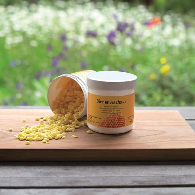 Bienenwachsdrops in einer Dose geschlossen und geöffnet zur Seite gekippt auf Holzbrett vor einer Wiese